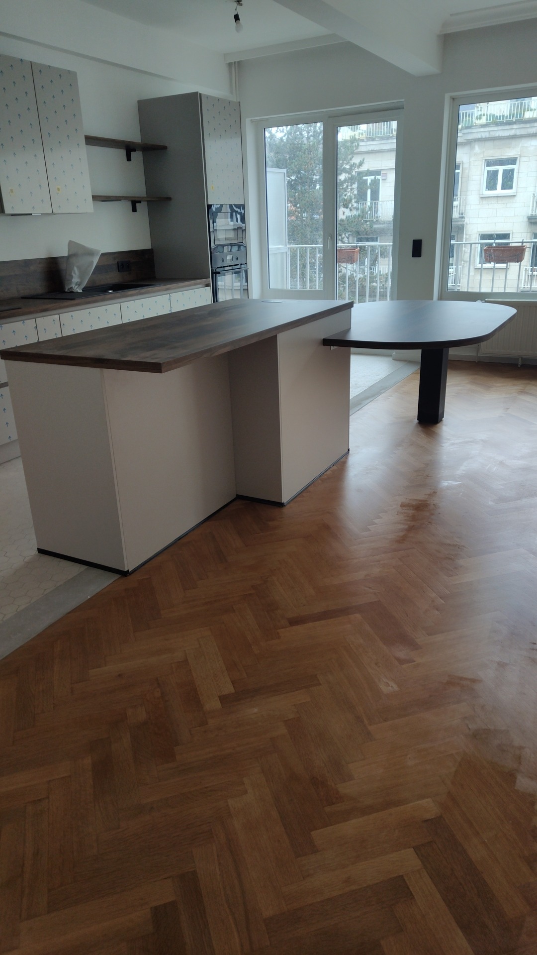 Wnętrze kuchni w trakcie wykończenia z wyspą kuchenną z blatem w kolorze ciemnego drewna, parkietem ułożonym w jodełkę i widokiem na budynek przez okno.