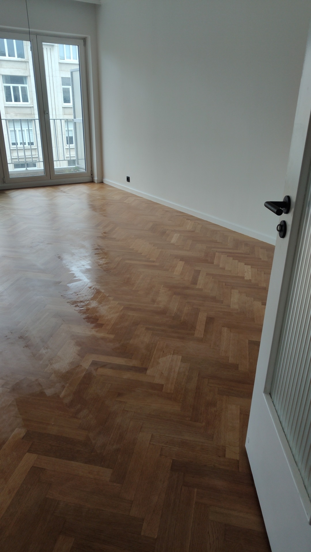 Jasne wnętrze z parkietem ułożonym w jodełkę, świeżo po lakierowaniu, z widocznym połyskiem na podłodze, białe ściany i fragment drzwi z czarną klamką.