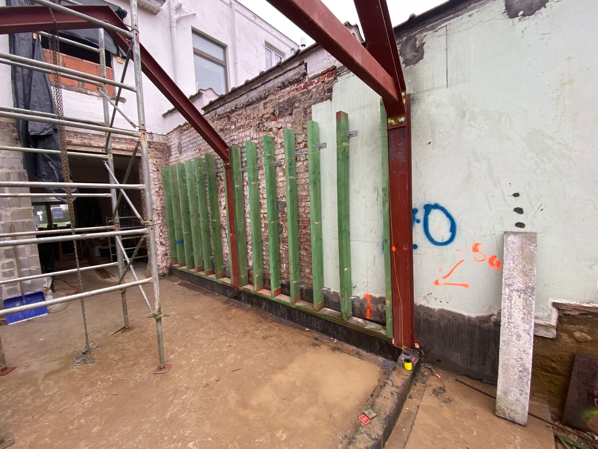 Konstrukcja nośna z belek stalowych i drewnianych słupków, przygotowana do murowania ściany na zewnątrz budynku z widocznymi elementami rusztowania i oznaczeniami na ścianie.