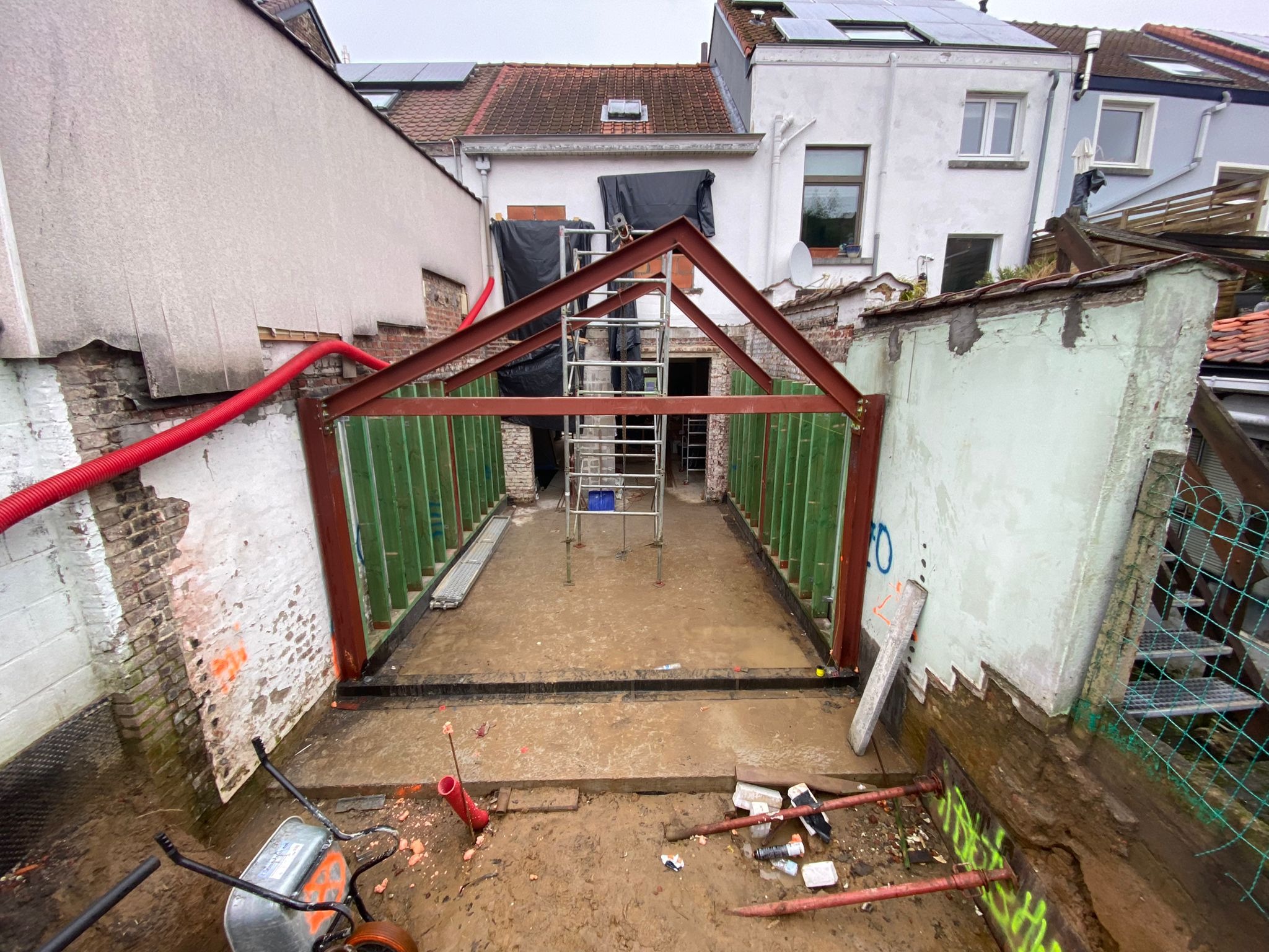 Widok na plac budowy podczas rozbudowy domu, z widoczną stalową konstrukcją szkieletową, szalunkiem i błotnistym podłożem. W tle widoczny budynek z panelami słonecznymi na dachu.