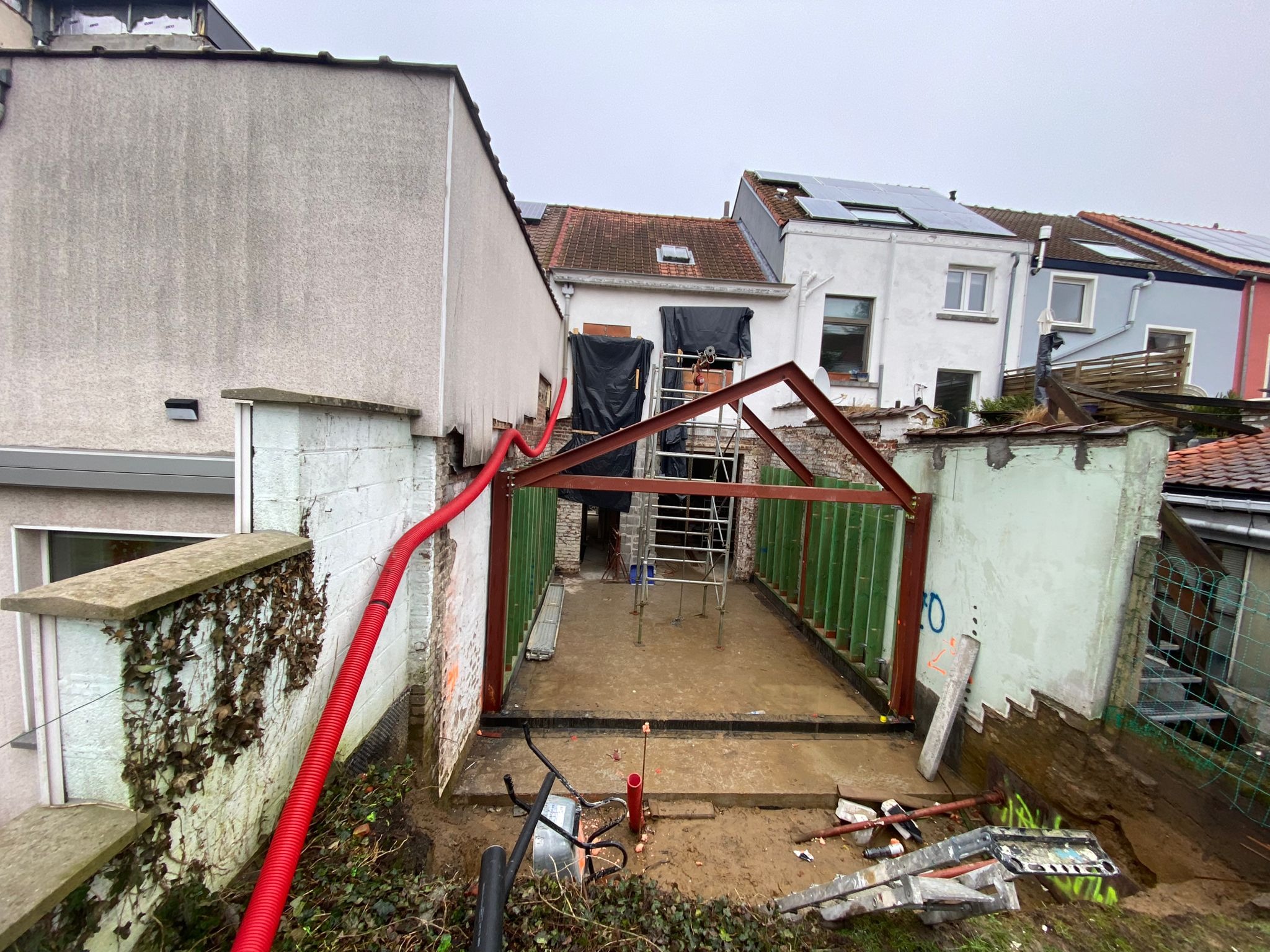 Budowa domu w trakcie realizacji: widok fundamentów z metalową konstrukcją szkieletową, czerwona rura instalacyjna biegnąca po ścianie budynku, drabina i narzędzia budowlane w tle.