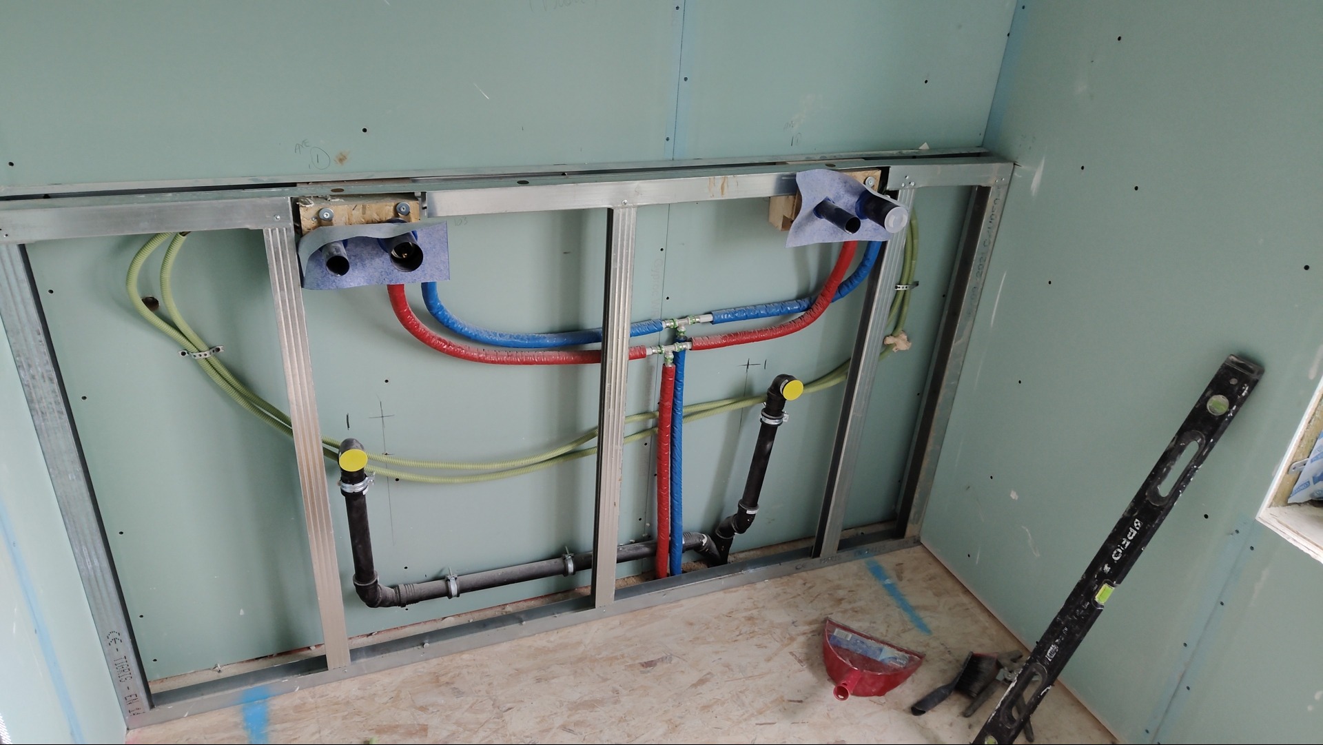 Wewnętrzna instalacja wodno-kanalizacyjna w łazience, widoczne rury PEX w kolorze czerwonym i niebieskim, rury kanalizacyjne z żółtymi zaślepkami, metalowy stelaż pod zabudowę z płyt karton-gips...