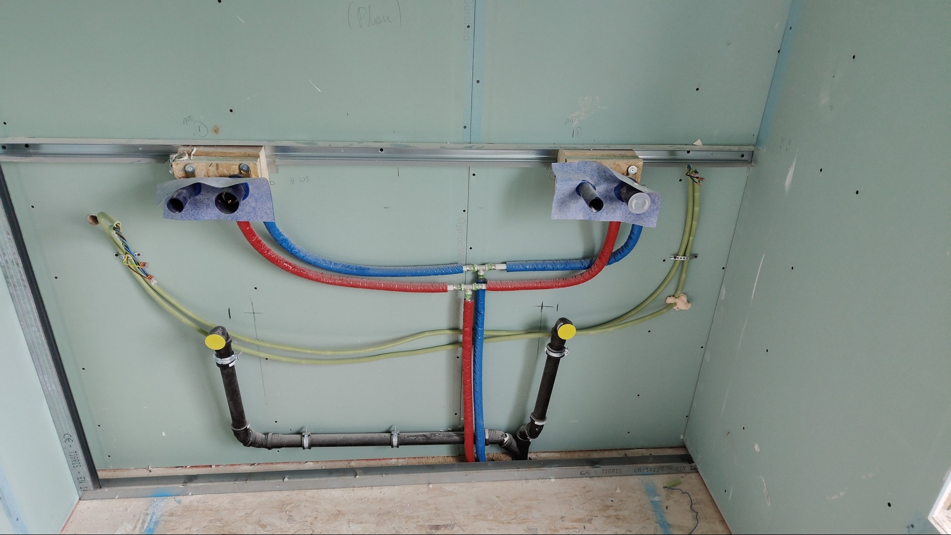 Ściana z płyt kartonowo-gipsowych w trakcie montażu z widocznymi instalacjami wodno-kanalizacyjnymi i elektrycznymi, rury w kolorze czerwonym i niebieskim, przewody żółte, czarne rury kanalizacyjne...