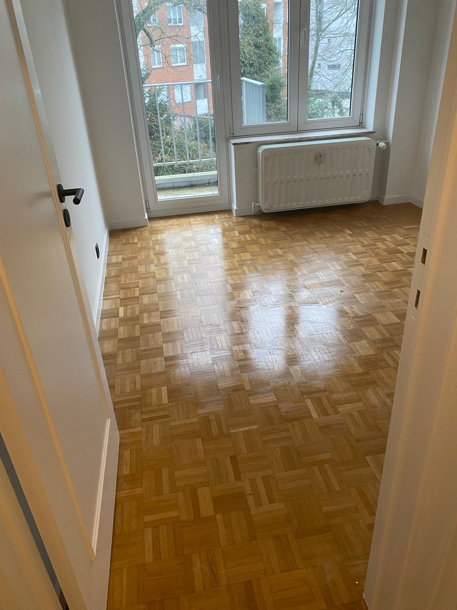 Pusty pokój z parkietem w jodełkę, białymi ścianami, grzejnikiem pod oknem balkonowym z widokiem na budynek mieszkalny.