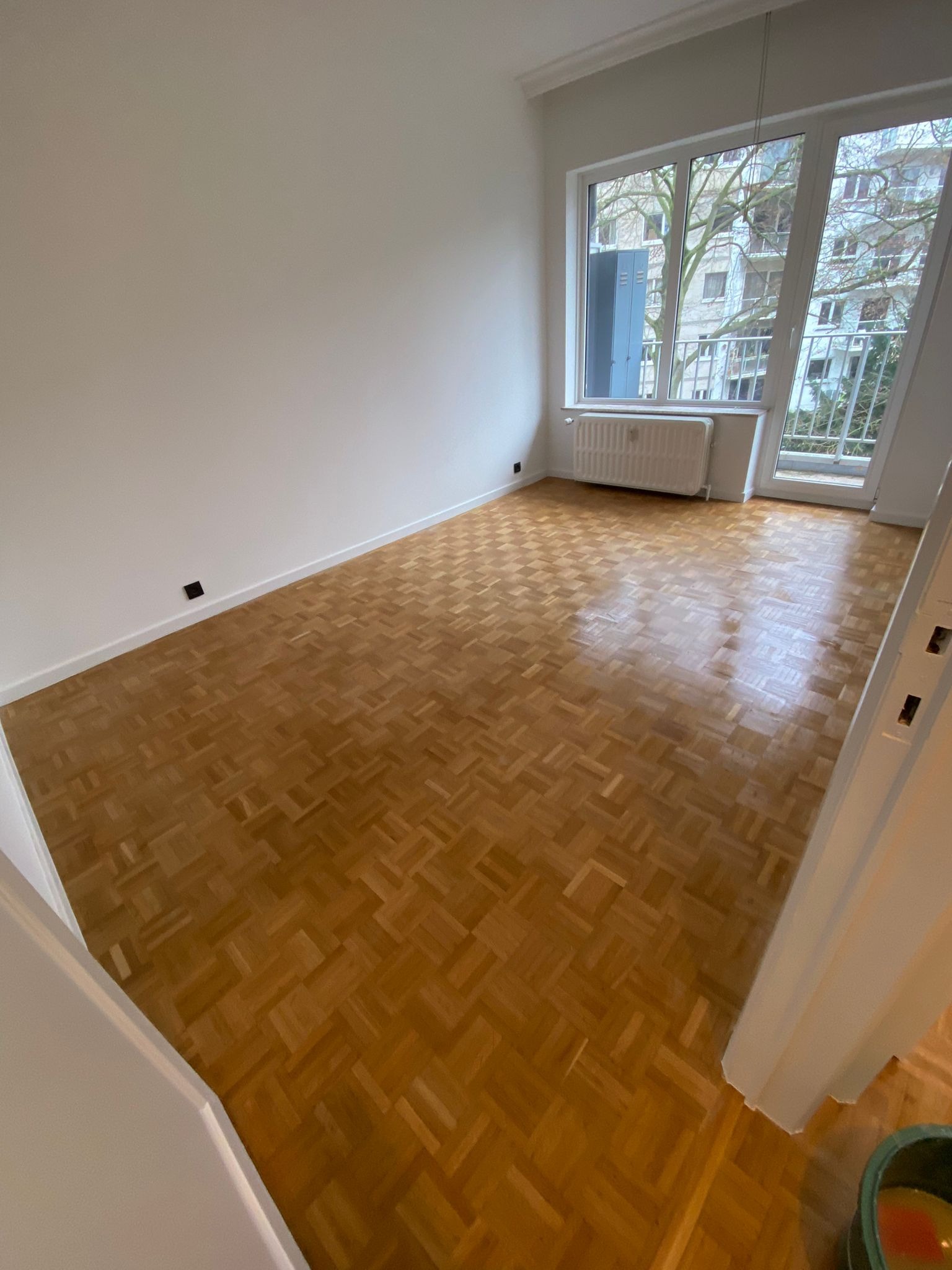 Pusty pokój z parkietem w jodełkę, świeżo pomalowanymi białymi ścianami i grzejnikiem pod oknem balkonowym z widokiem na budynek mieszkalny.