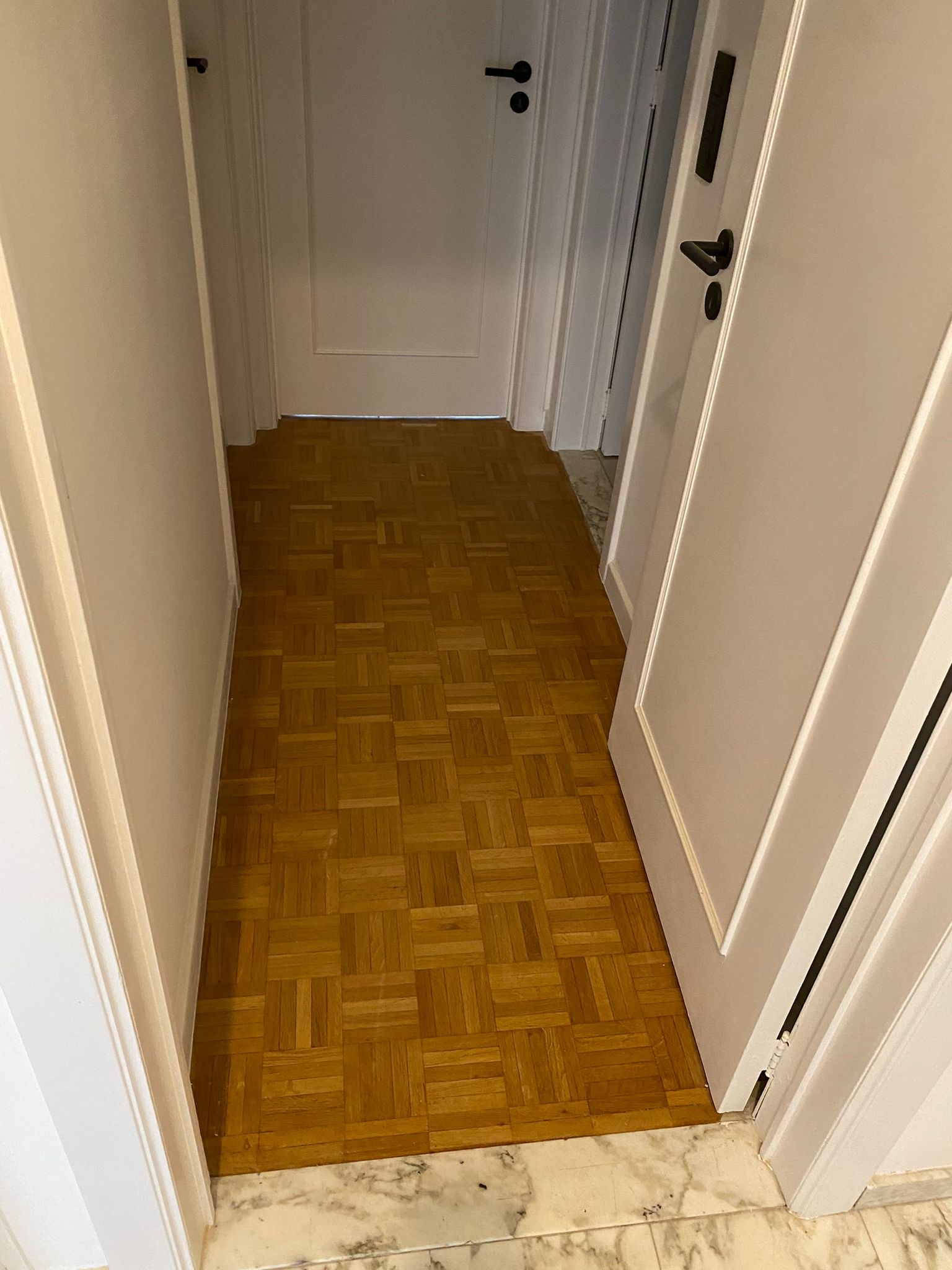 Korytarz z parkietem ułożonym w kwadraty, widoczne białe drzwi z czarnymi klamkami, marmurowy cokół przy podłodze.