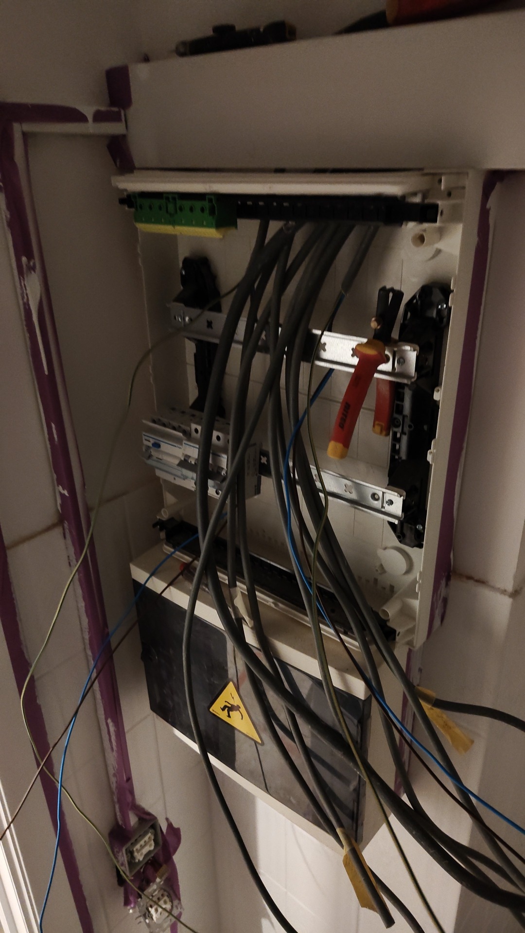 Rozdzielnica elektryczna z licznymi przewodami, narzędzia (kombinerki) wewnątrz, widoczny znak ostrzegawczy o wysokim napięciu, instalacja w trakcie prac na tle białych płytek.