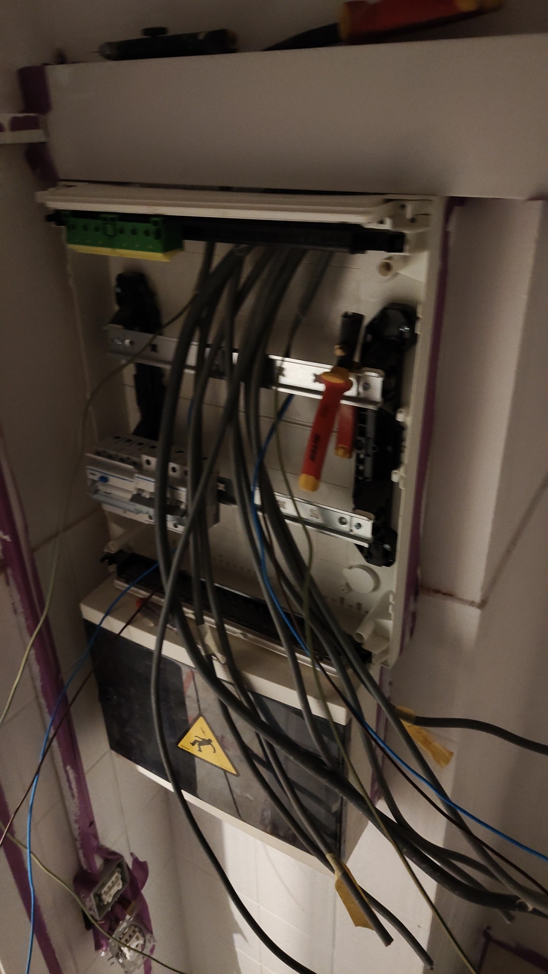 Otwarta skrzynka elektryczna z licznymi kablami, zabezpieczeniami i narzędziami, widoczny znak ostrzegawczy o ryzyku porażenia prądem, instalacja w trakcie prac.