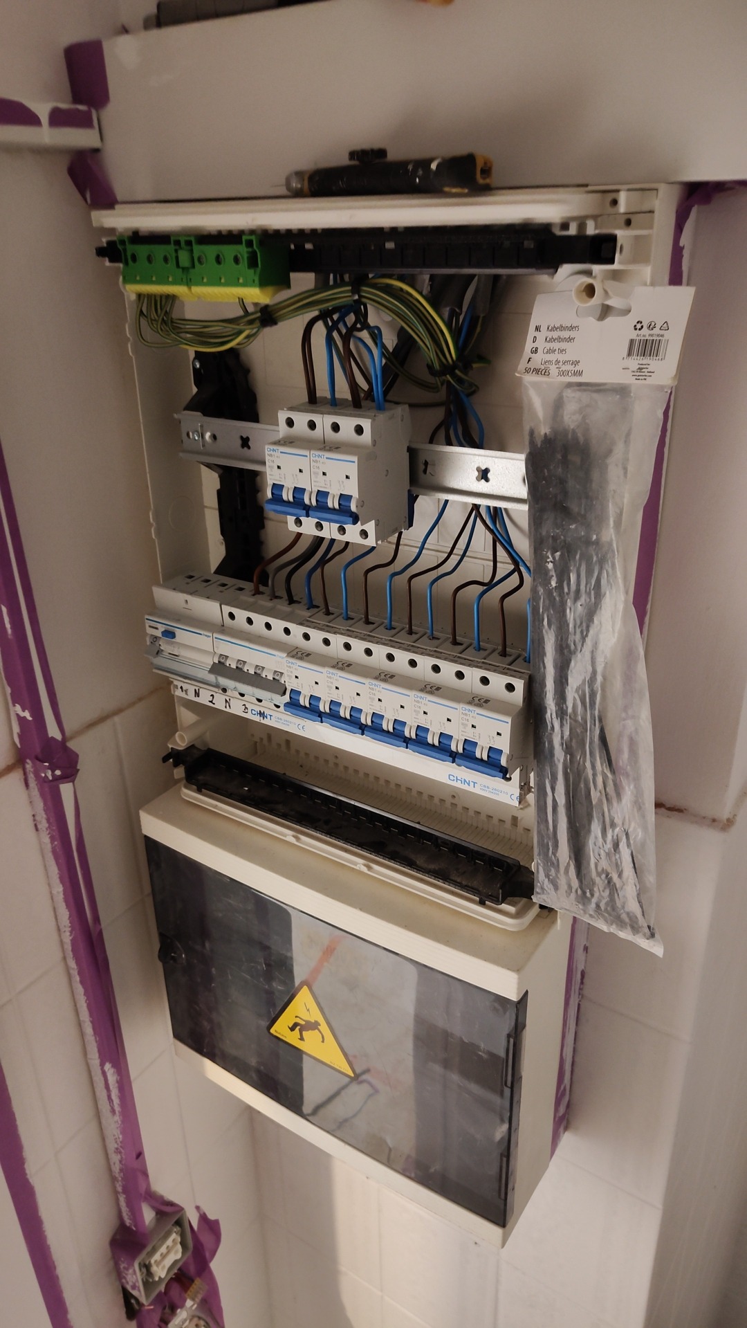 Otwarta skrzynka rozdzielcza z zabezpieczeniami, przewodami, listwami zaciskowymi i paczką opasek kablowych, zamontowana na białej ścianie z fioletowymi elementami dekoracyjnymi.
