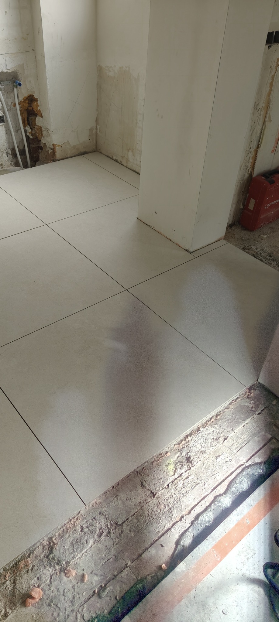 Wyłożona jasnymi płytkami podłoga w trakcie remontu, widoczne fragmenty niezakończonych prac przy ścianach i progu. Czerwona skrzynka narzędziowa w tle.