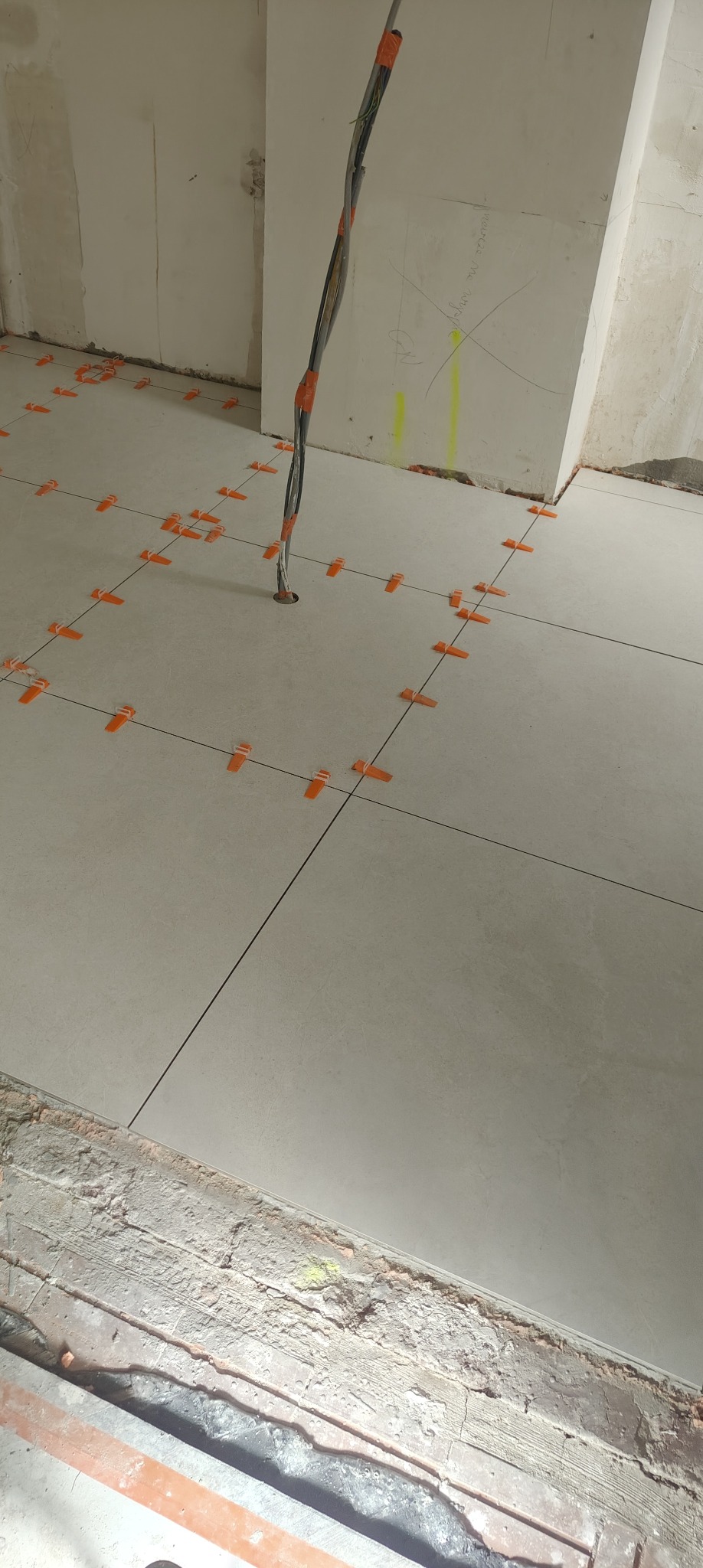 Układanie jasnych płytek podłogowych z użyciem pomarańczowych klipsów poziomujących. W tle widoczne kable elektryczne i surowe ściany w trakcie remontu.