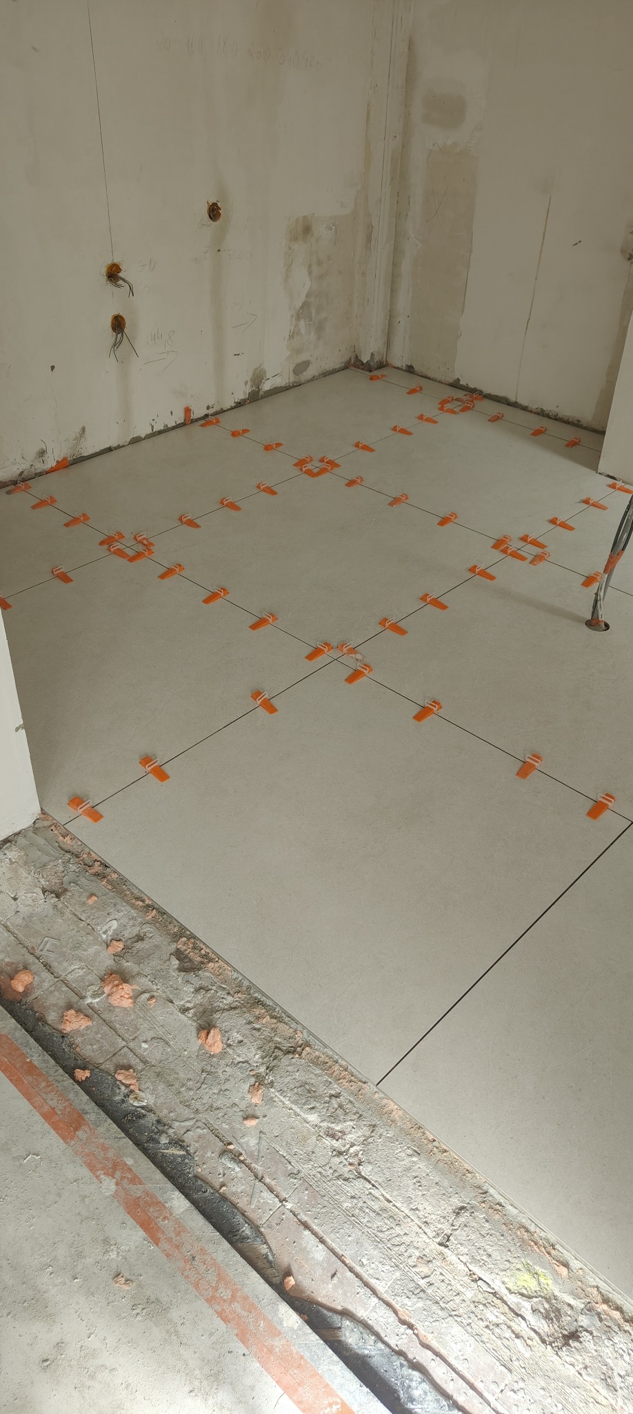 Układanie jasnych płytek podłogowych z użyciem pomarańczowych klipsów poziomujących. Widoczne linie fug i surowe ściany w tle. Etap prac wykończeniowych.