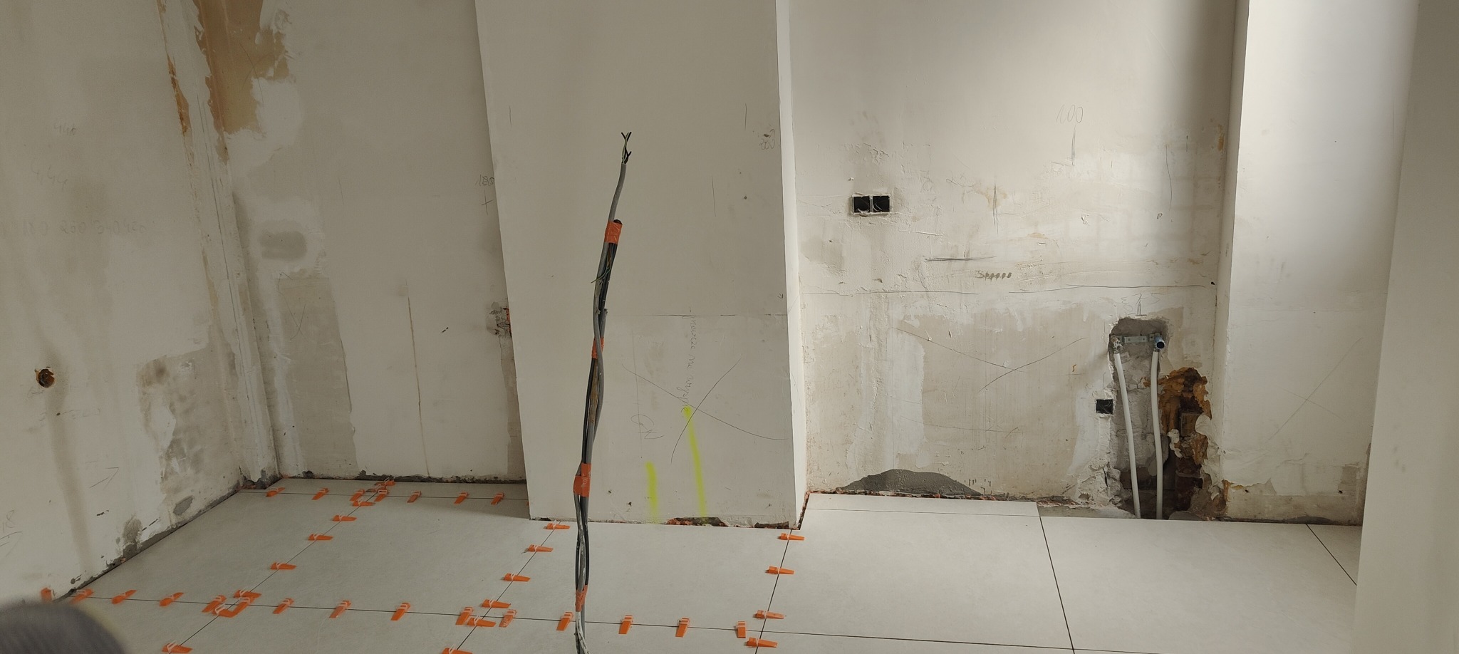 Układanie białych płytek podłogowych w pomieszczeniu w trakcie remontu. Widoczne kable elektryczne i ślady po instalacjach na ścianach.