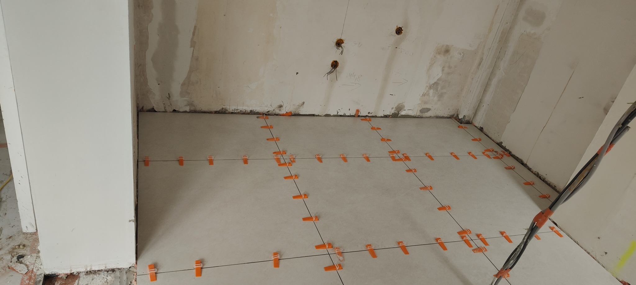 Układanie jasnych płytek podłogowych z użyciem pomarańczowych krzyżyków dystansowych i czarnych sznurków. Widoczne wyprowadzenia elektryczne na ścianie.