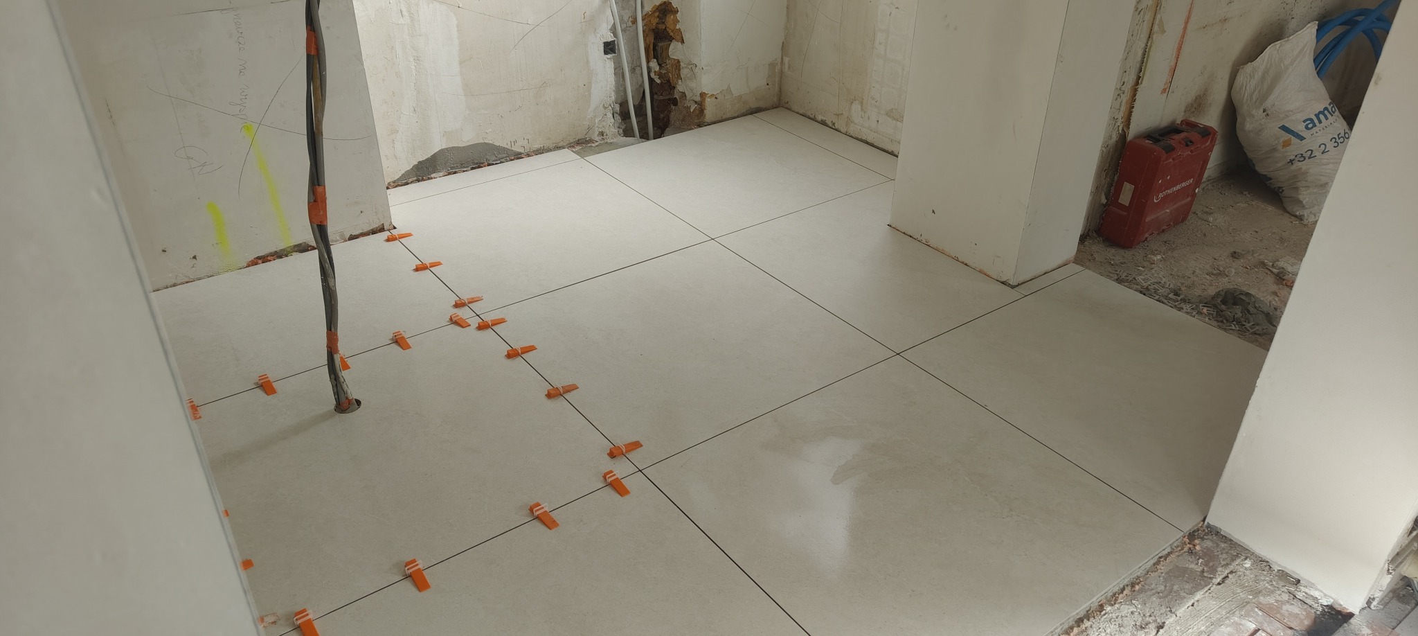 Ułożone jasne płytki podłogowe z pomarańczowymi krzyżykami dystansowymi, widoczne przewody elektryczne i przygotowane ściany do dalszych prac wykończeniowych.
