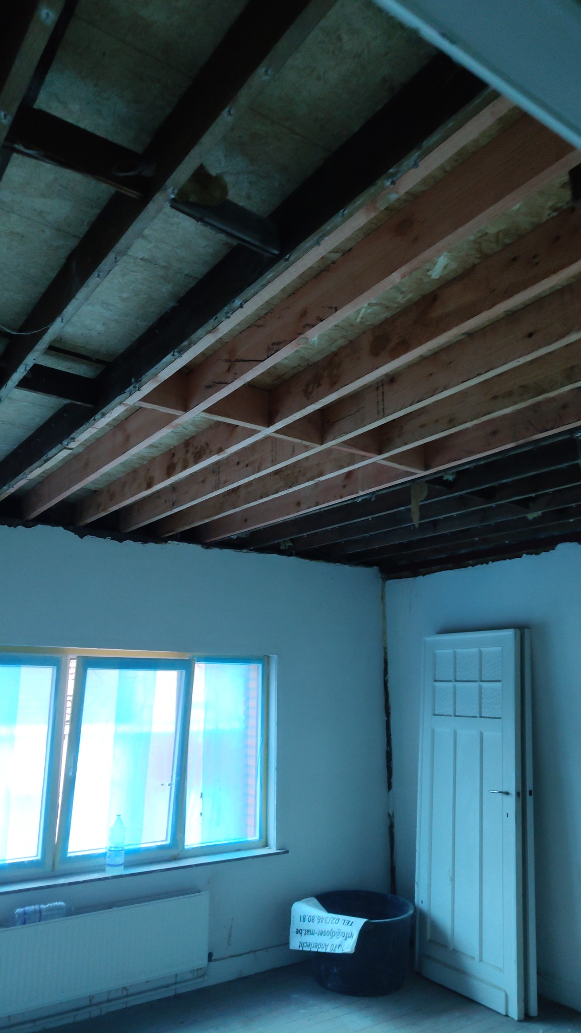 Montaż drewnianej konstrukcji sufitu w Sępólnie Krajeńskim. Widoczne belki i płyty OSB. W tle okno z niebieską folią i białe ściany. Prace remontowe w trakcie.