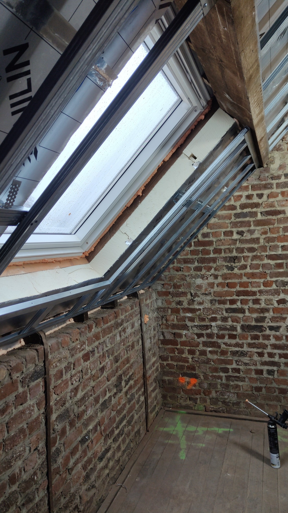 Montaż okna dachowego w surowym wnętrzu z ceglanymi ścianami i drewnianą podłogą. Widoczne metalowe profile i pianka montażowa. Oznakowania na podłodze.