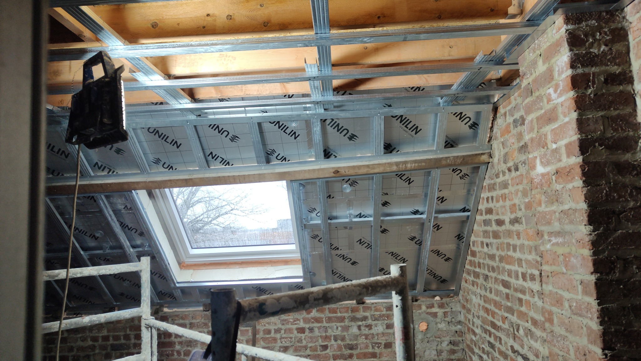 Montaż izolacji poddasza z oknem dachowym, widoczne metalowe profile i materiał izolacyjny z nadrukiem. Fragment rusztowania i ściana z cegły w surowym stanie.