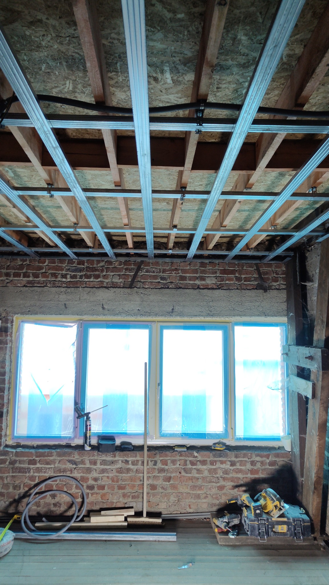 Montaż metalowej konstrukcji sufitu podwieszanego w pomieszczeniu z ceglaną ścianą i oknami zabezpieczonymi folią. Widoczne narzędzia i materiały budowlane.