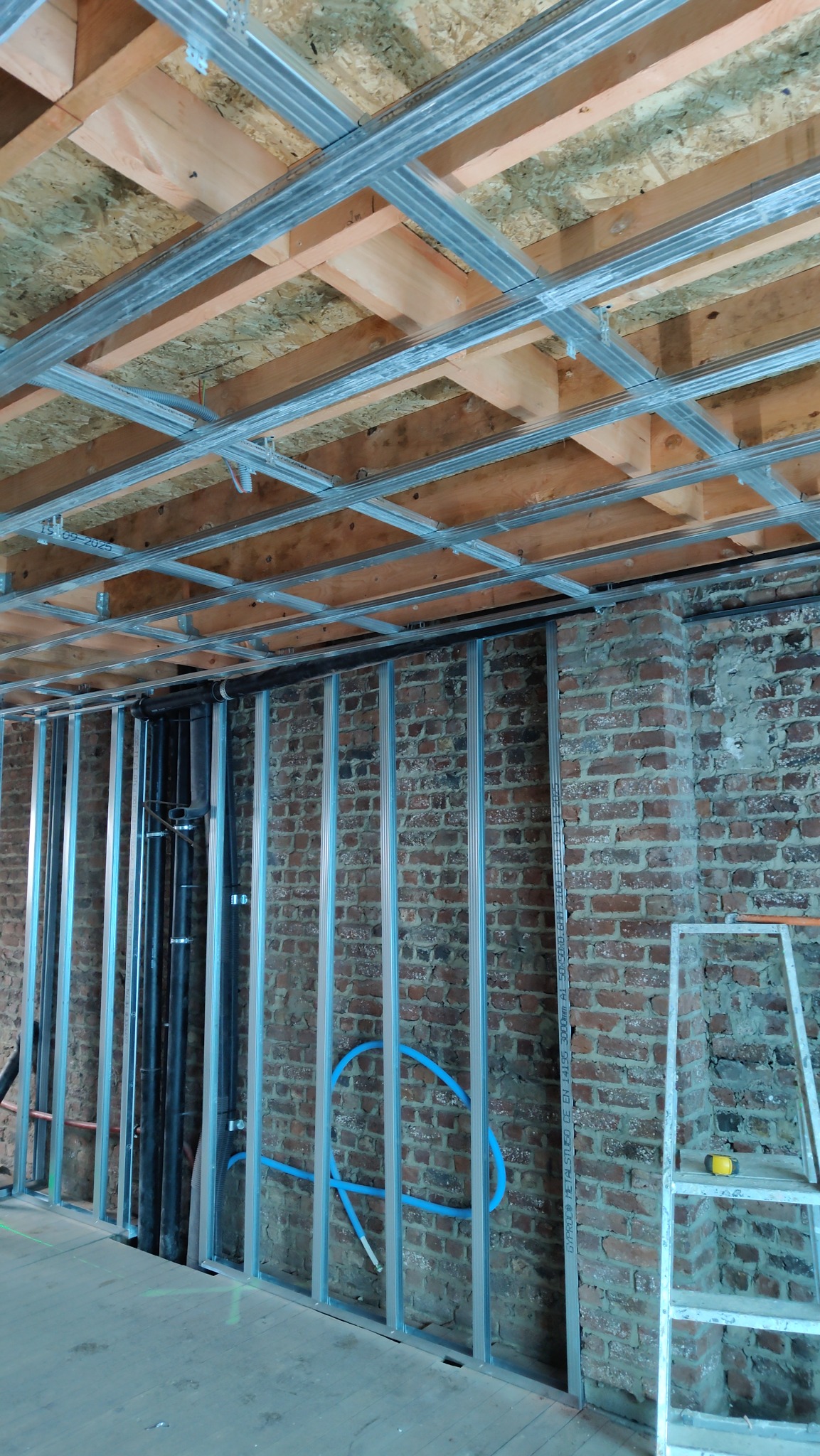 Montaż metalowej konstrukcji sufitu podwieszanego i ścian działowych na tle ceglanej ściany. Widoczne instalacje. Etap prac wykończeniowych w surowym wnętrzu.