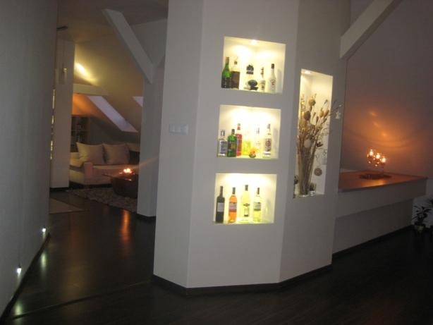 Wnętrze domu z wnękami w ścianie, podświetlone półki z dekoracjami i butelkami, widok na salon z kanapą i oknami dachowymi.