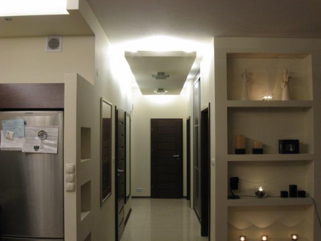 Wykończony korytarz z podwieszanym sufitem z oświetleniem punktowym, wbudowanymi półkami dekoracyjnymi z oświetleniem i lustrami na ścianach, widoczna lodówka ze stali nierdzewnej.