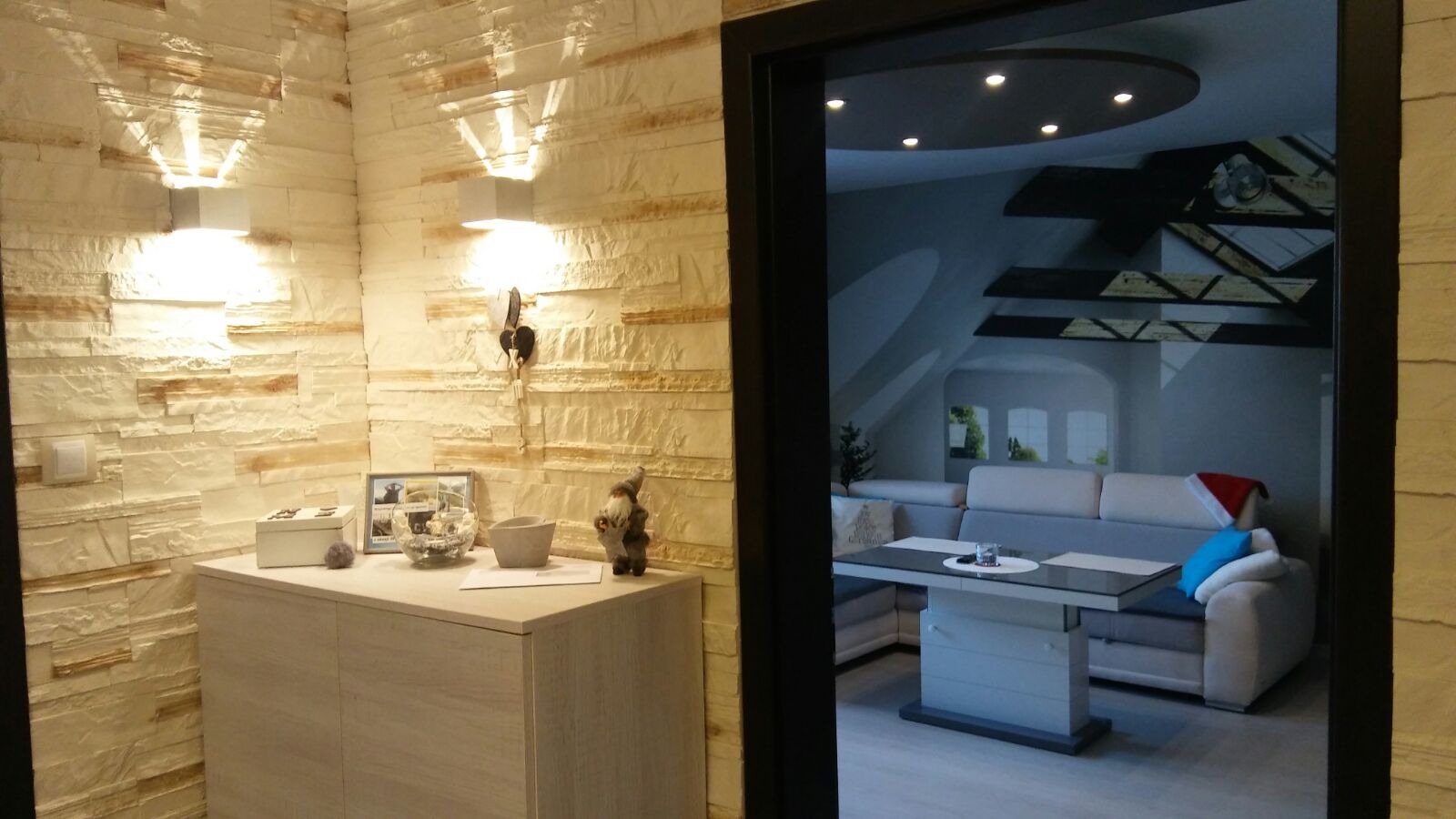 Salon po remoncie: ściana z dekoracyjnej cegły z oświetleniem punktowym, szafka z dekoracjami, widok na salon z narożnikiem i dekoracją sufitu.