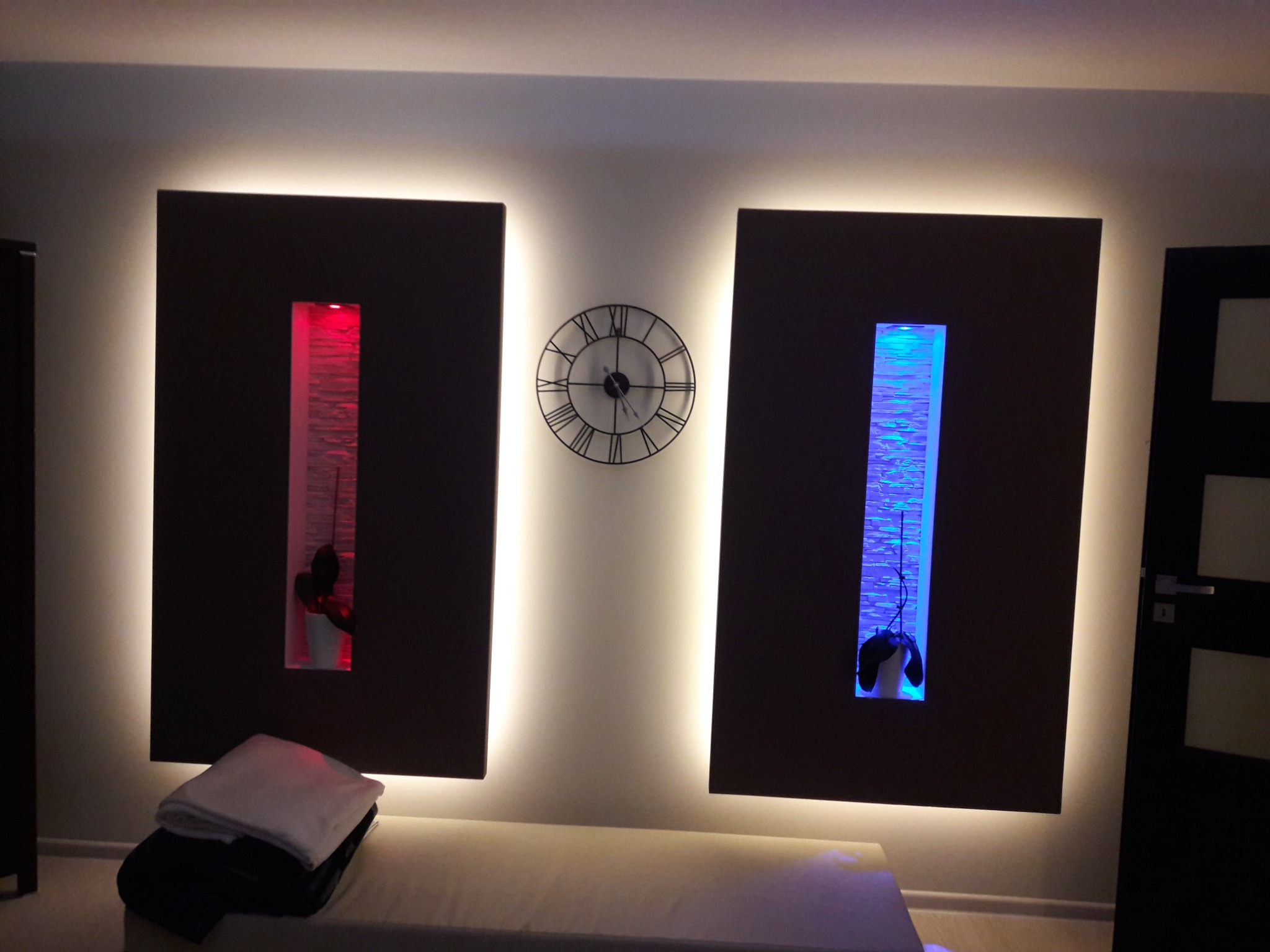 Nowoczesna aranżacja ściany w Szczecinie z dwoma podświetlonymi panelami dekoracyjnymi, wnękami z czerwonym i niebieskim oświetleniem oraz zegarem na ścianie