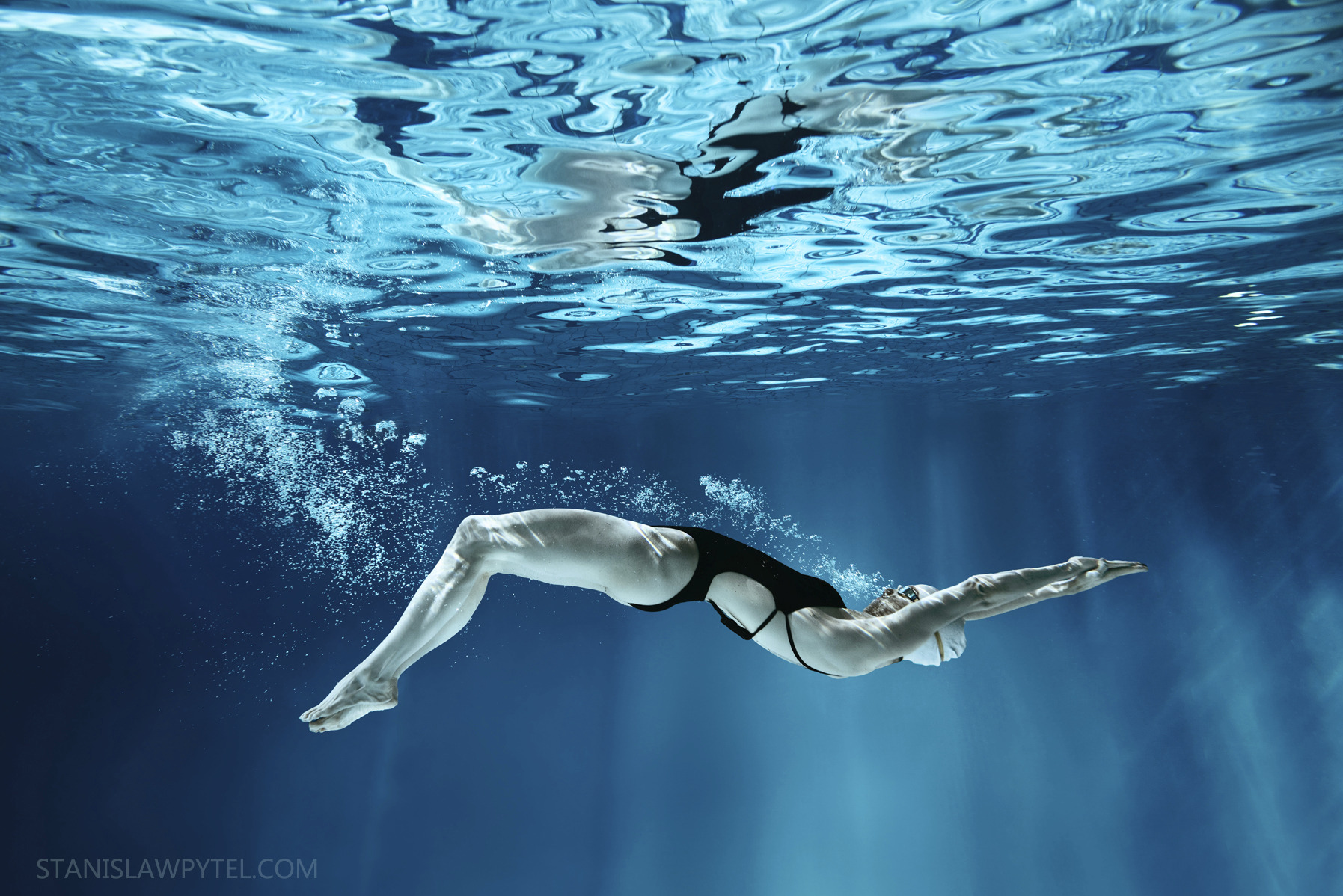 Kobieta w czarnym stroju kąpielowym płynie pod wodą, widoczna z dołu, z bąbelkami powietrza i odbiciem na powierzchni wody.