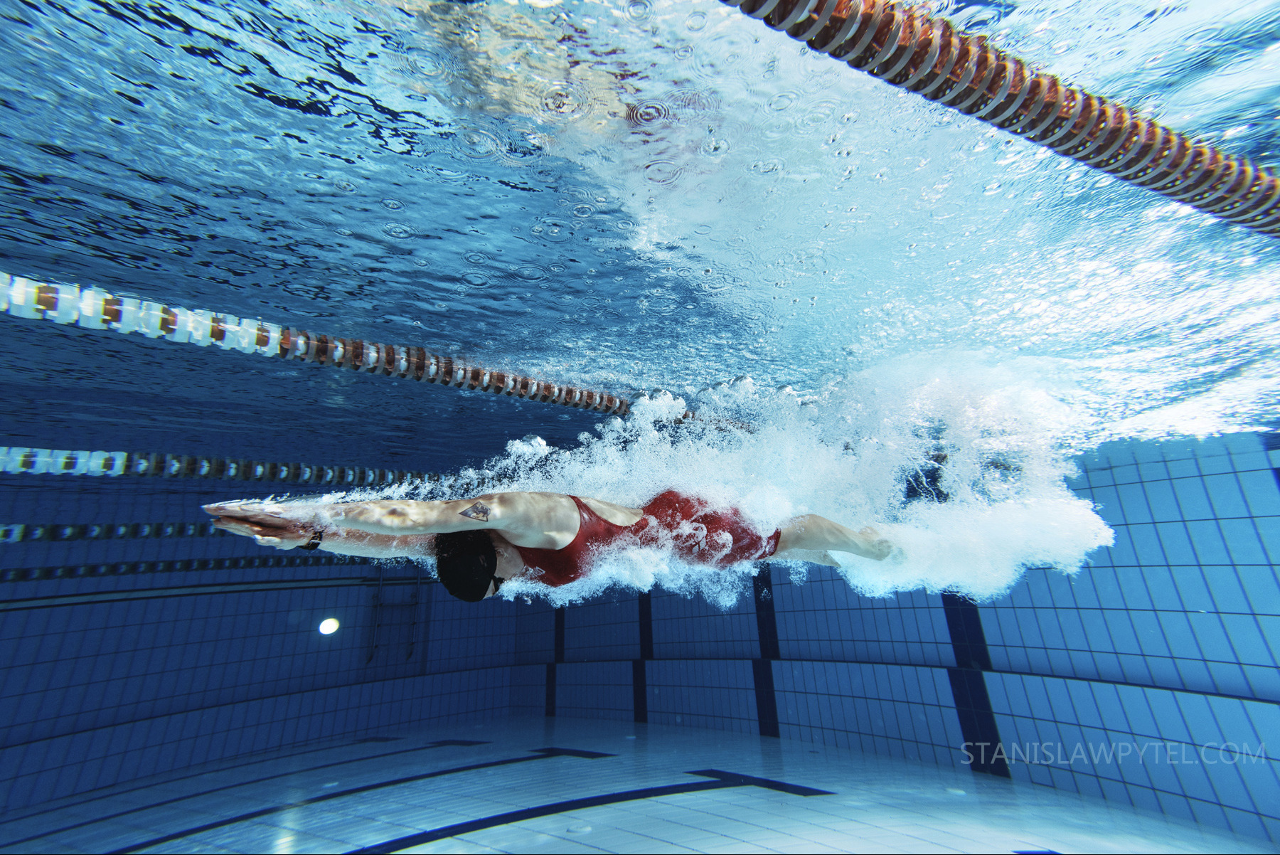 Podwodny widok pływaczki w czerwonym stroju, płynącej w basenie sportowym, z widocznymi torami i bąbelkami powietrza.