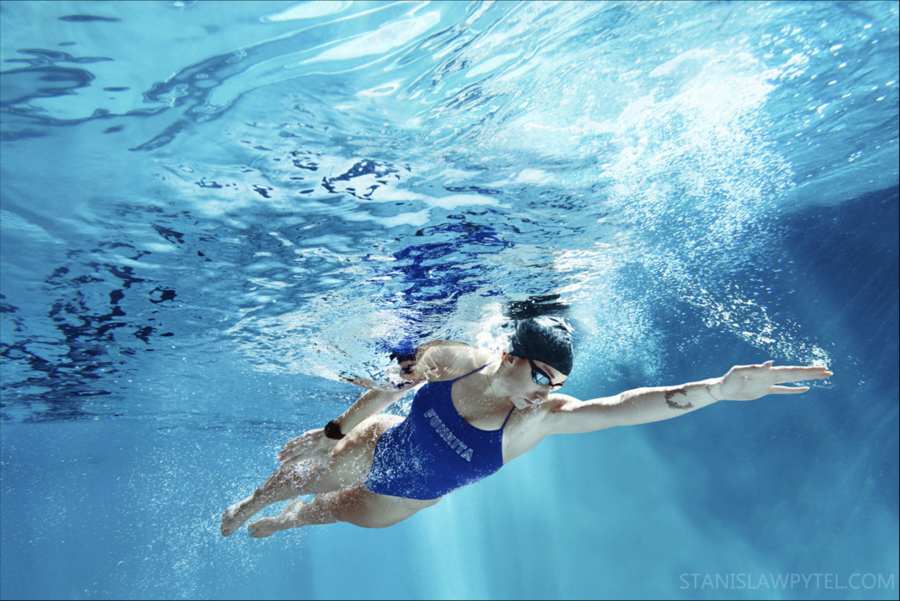 Kobieta w czepku i okularach pływackich płynie kraulem pod wodą w basenie, widok z dołu, bąbelki powietrza, woda w odcieniach błękitu.