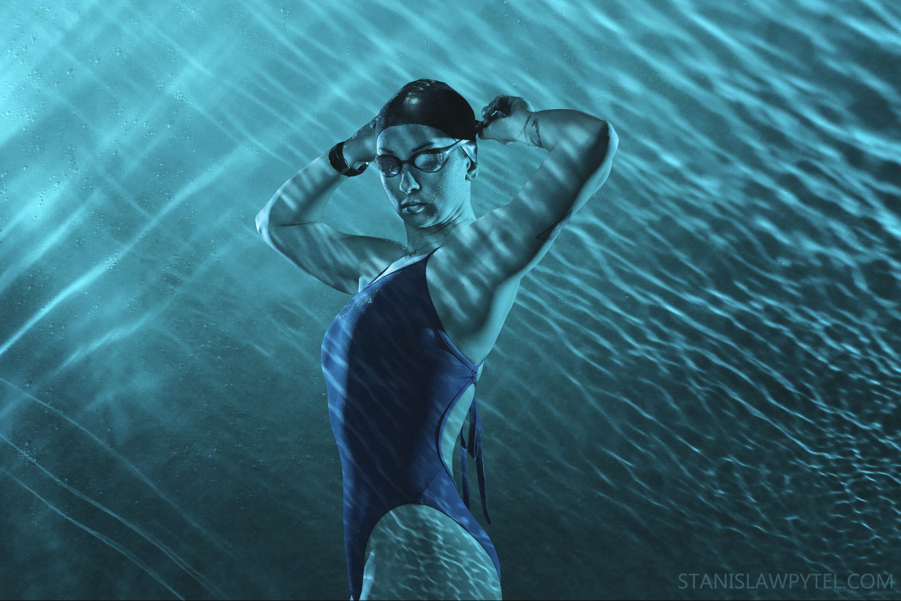 Kobieta w stroju kąpielowym, czepku i okularach pływackich, poprawia czepka na tle turkusowej wody z refleksami światła.