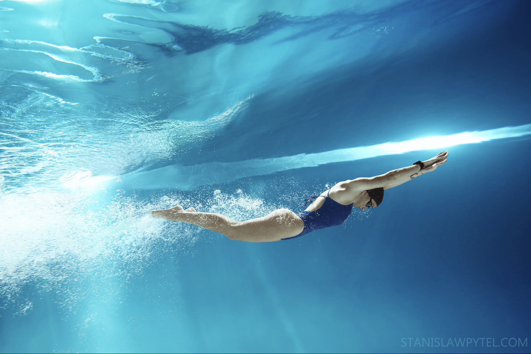 Kobieta w niebieskim stroju kąpielowym płynie pod wodą w basenie, wykonując dynamiczny ruch, widoczne refleksy światła na powierzchni wody i bąbelki powietrza.