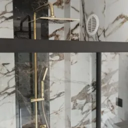 Złota armatura prysznicowa, deszczownica i bateria, w kabinie wyłożonej marmurowymi płytkami w odcieniach bieli, brązu i czerni, widoczny fragment wentylacji.
