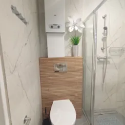 Nowoczesna łazienka z kabiną prysznicową, podwieszaną toaletą i drewnianą zabudową. Białe marmurowe płytki, szara podłoga, minimalistyczny design.