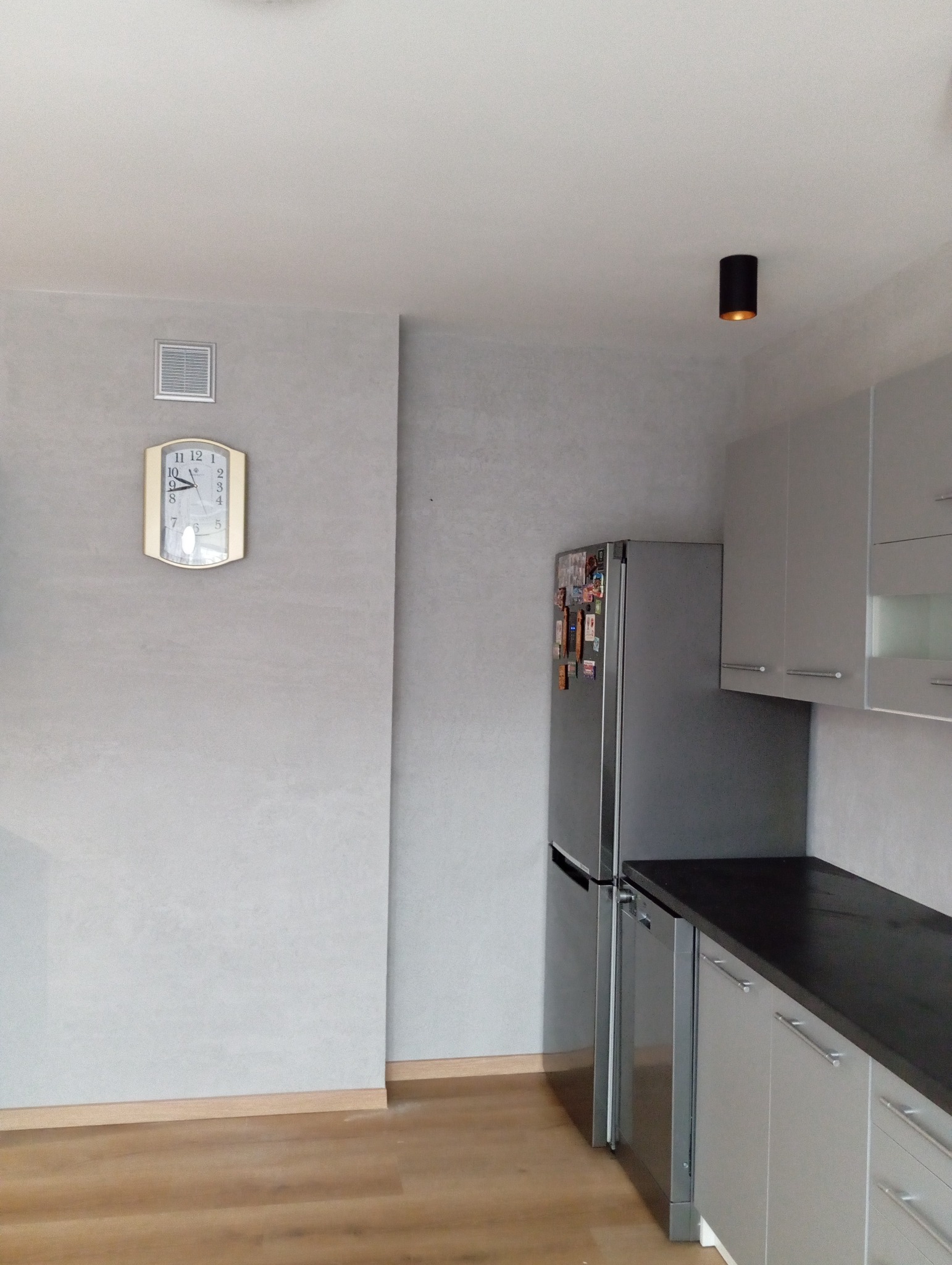 Szara kuchnia z zegarem ściennym, lodówką z magnesami i czarnym punktowym oświetleniem sufitowym.