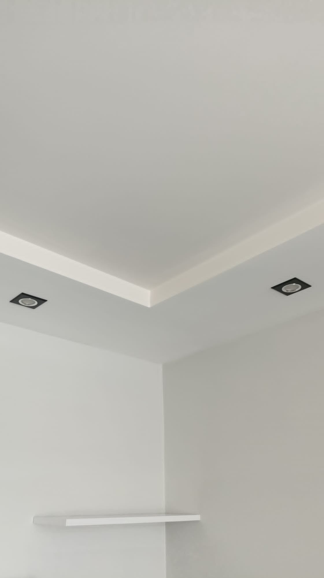 Białe wnętrze z minimalistycznym oświetleniem punktowym w suficie podwieszanym oraz małą półką przy ścianie.