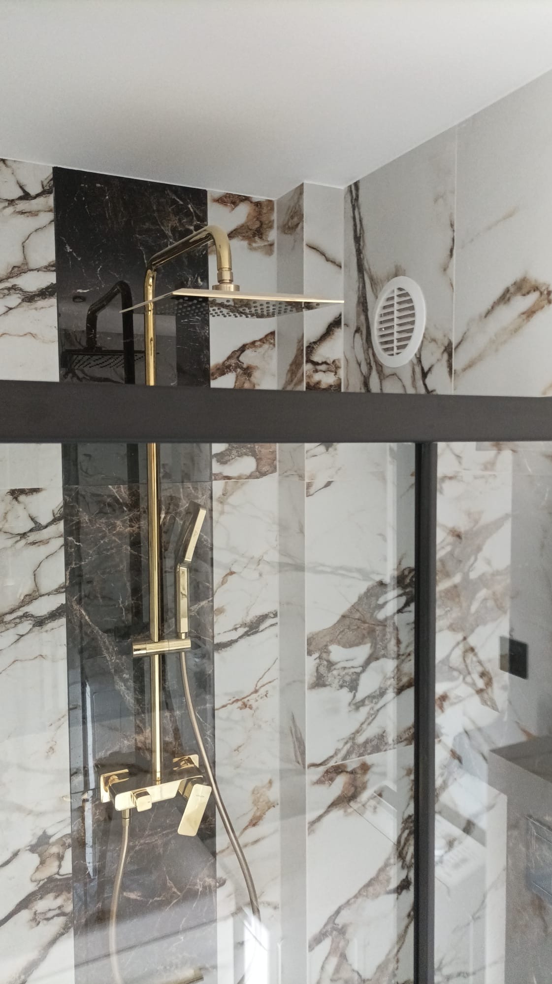 Złota armatura prysznicowa, deszczownica i bateria, w kabinie wyłożonej marmurowymi płytkami w odcieniach bieli, brązu i czerni, widoczny fragment wentylacji.