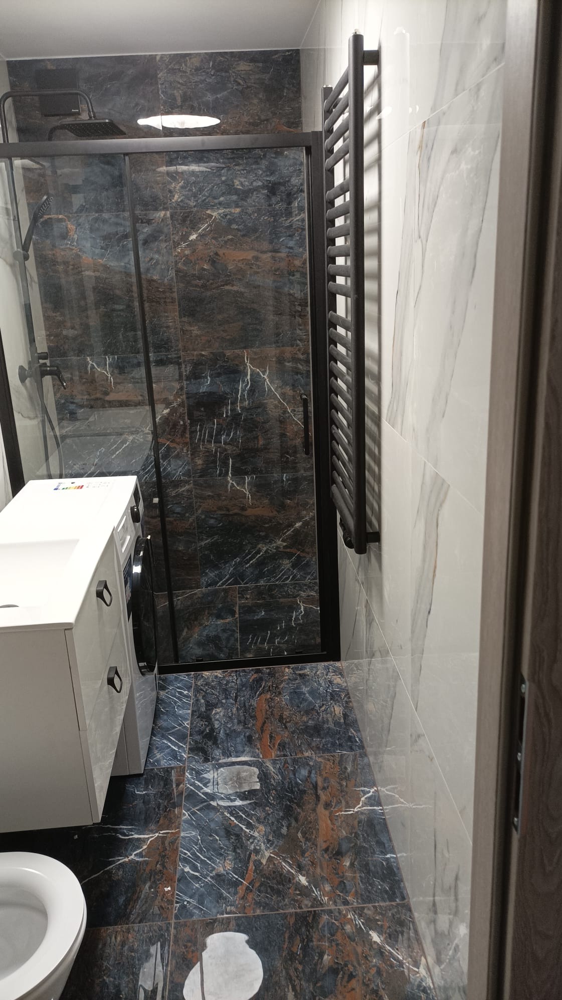 Nowoczesna łazienka z kabiną prysznicową typu walk-in, grzejnikiem drabinkowym w kolorze antracytowym i płytkami imitującymi marmur w odcieniach szarości, brązu i bieli.