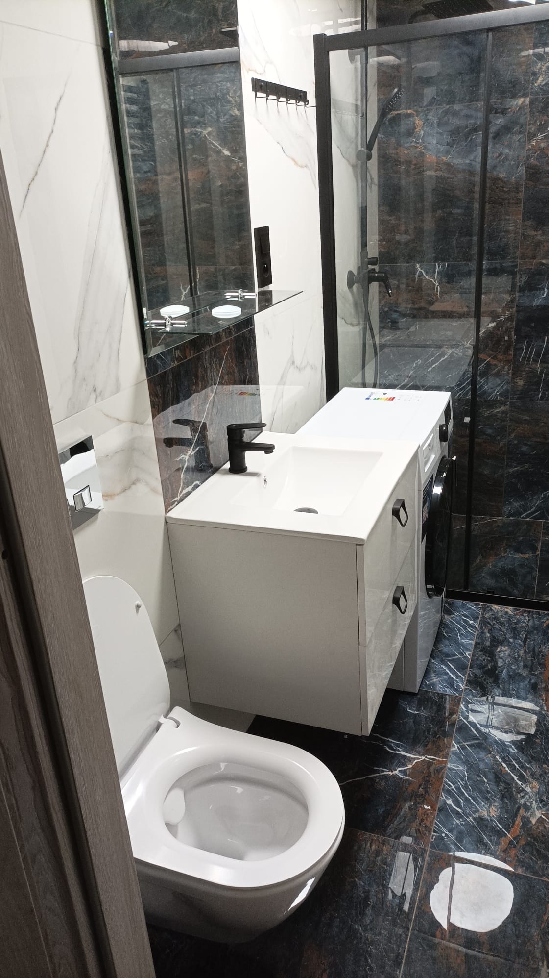 Nowoczesna łazienka z marmurową podłogą i ścianami, białą umywalką z czarną baterią, toaletą podwieszaną i kabiną prysznicową z czarną ramą, obok umywalki pralka w zabudowie.