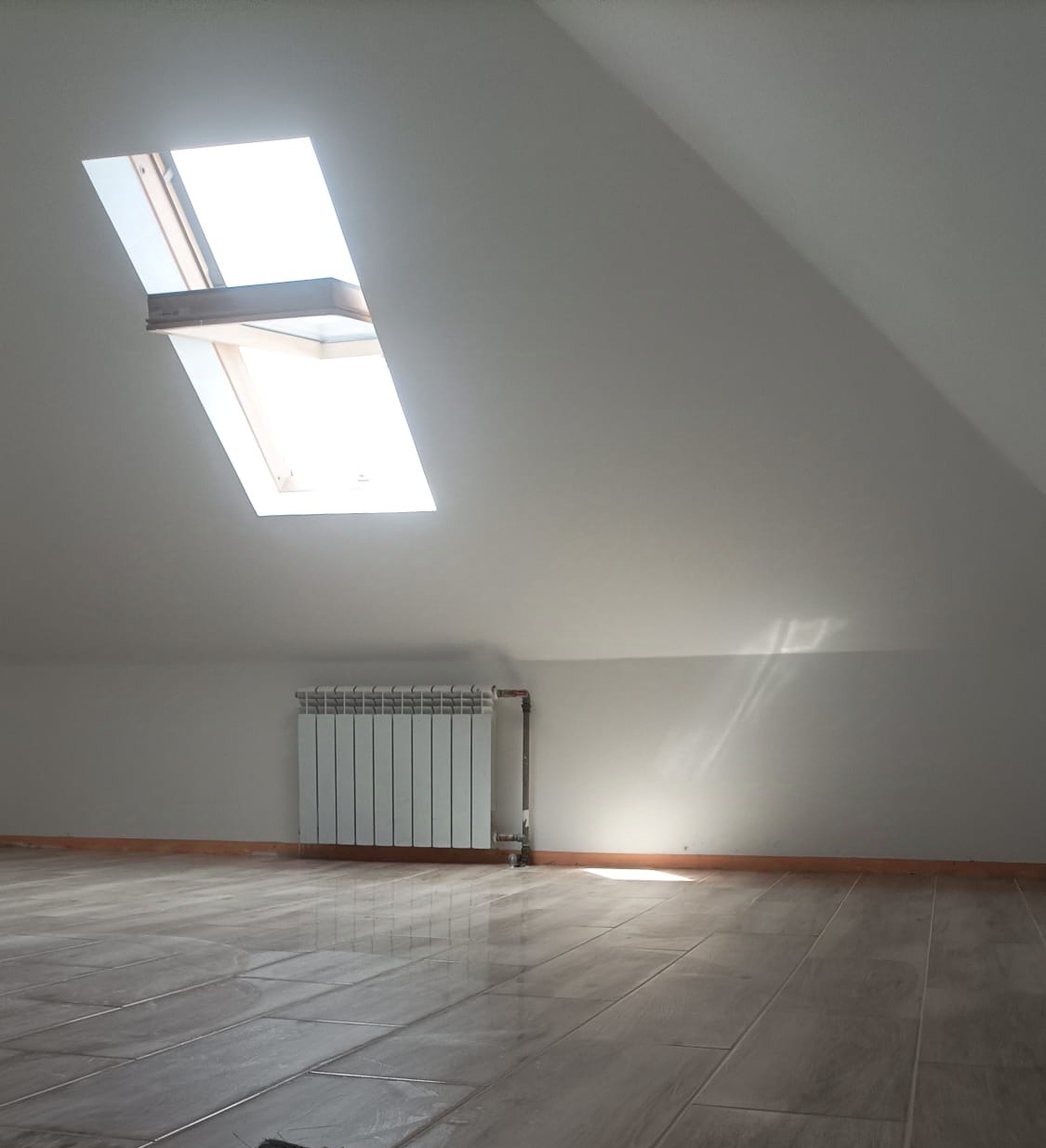 Poddasze z oknem dachowym, grzejnikiem i błyszczącą, ułożoną pod kątem podłogą z płytek imitujących drewno, oświetlone naturalnym światłem.