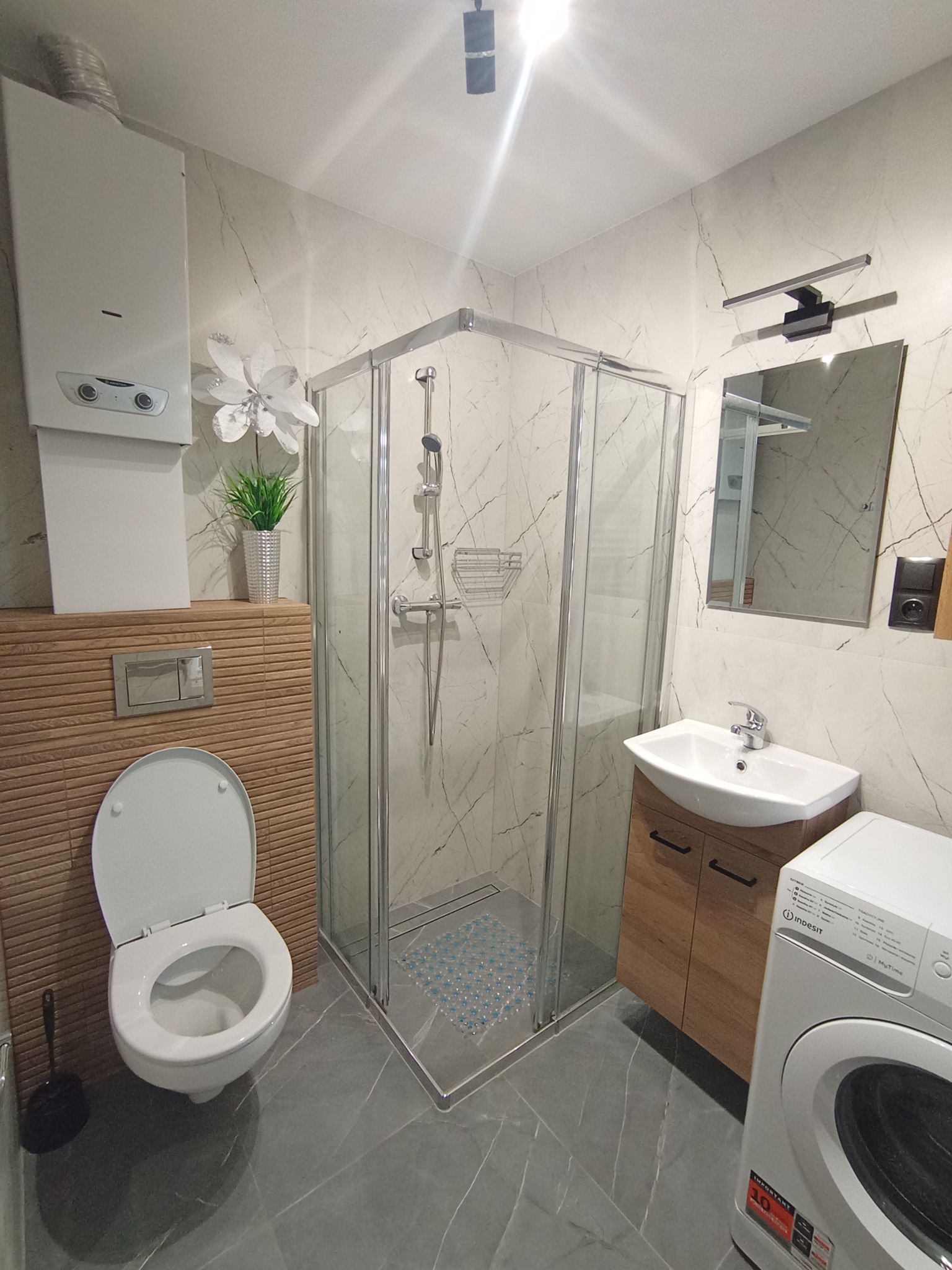 Wykończona łazienka z kabiną prysznicową, umywalką z szafką, toaletą i pralką. Ściany wyłożone marmurowymi płytkami, szare płytki podłogowe z białymi żyłami.