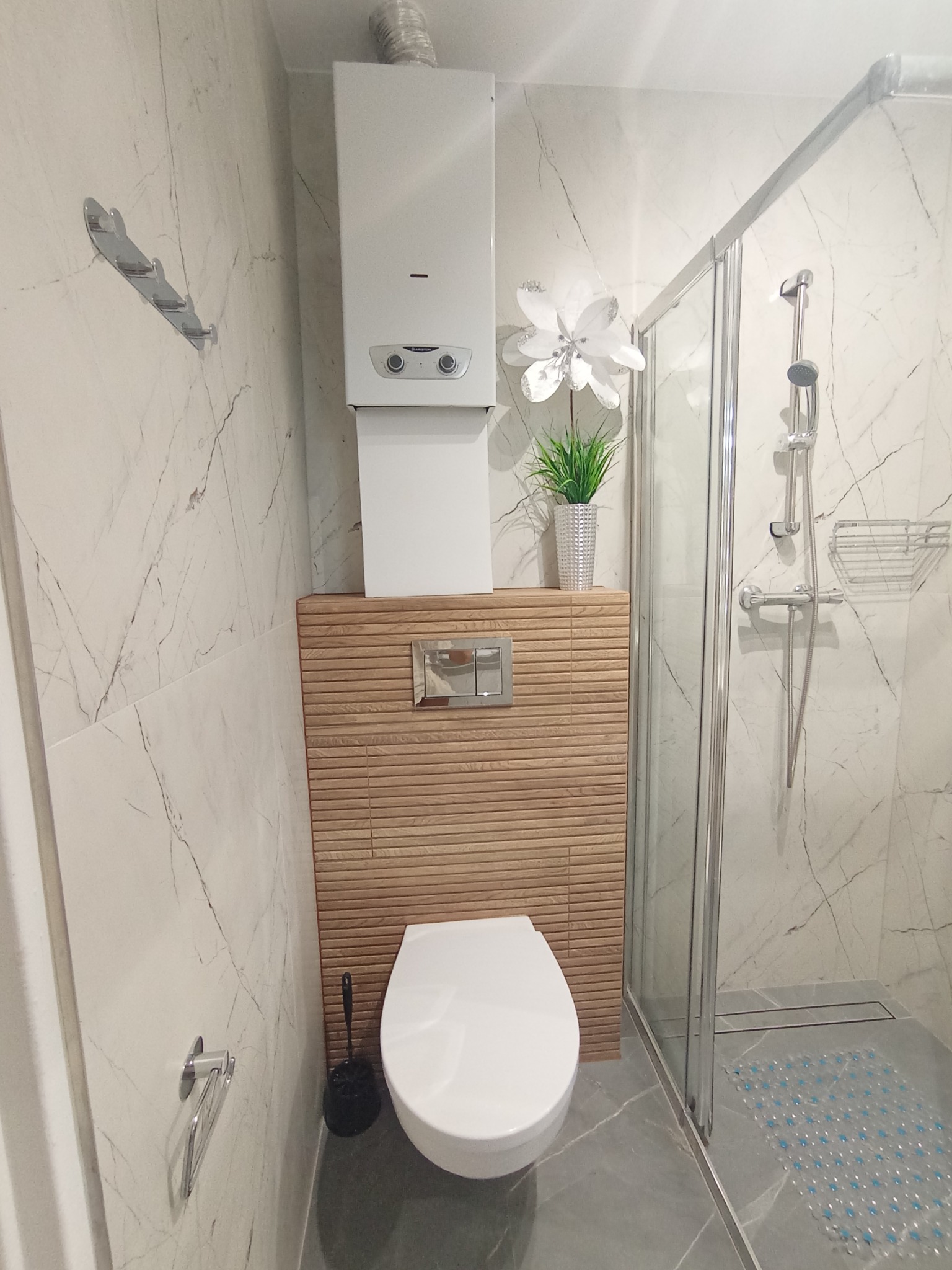 Nowoczesna łazienka z kabiną prysznicową, podwieszaną toaletą i drewnianą zabudową. Białe marmurowe płytki, szara podłoga, minimalistyczny design.