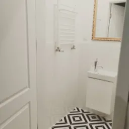 Nowoczesna, biała łazienka z geometryczną, czarno-białą podłogą, wiszącą umywalką i grzejnikiem drabinkowym, dopełniona lustrem w złotej ramie.