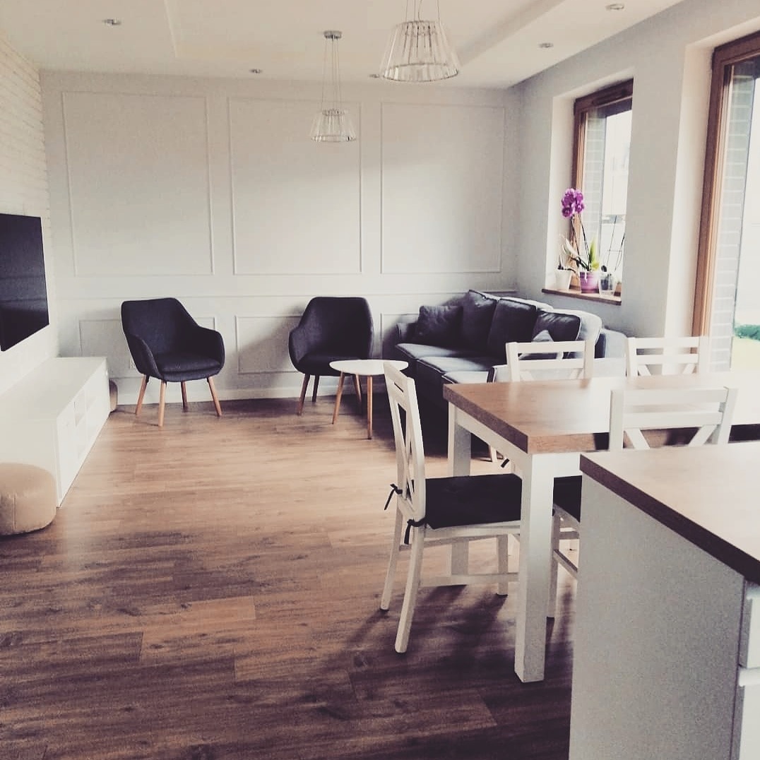 Jasne, przestronne wnętrze salonu z drewnianą podłogą, szarą sofą i krzesłami, białymi panelami ściennymi oraz nowoczesnym oświetleniem wiszącym.