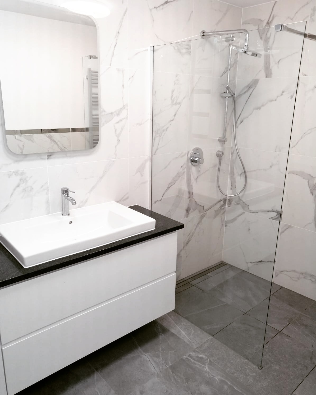 Nowoczesna łazienka z kabiną prysznicową typu walk-in, umywalką nablatową na białej szafce z czarnym blatem i lustrem z zaokrąglonymi rogami, wykończona płytkami imitującymi marmur.