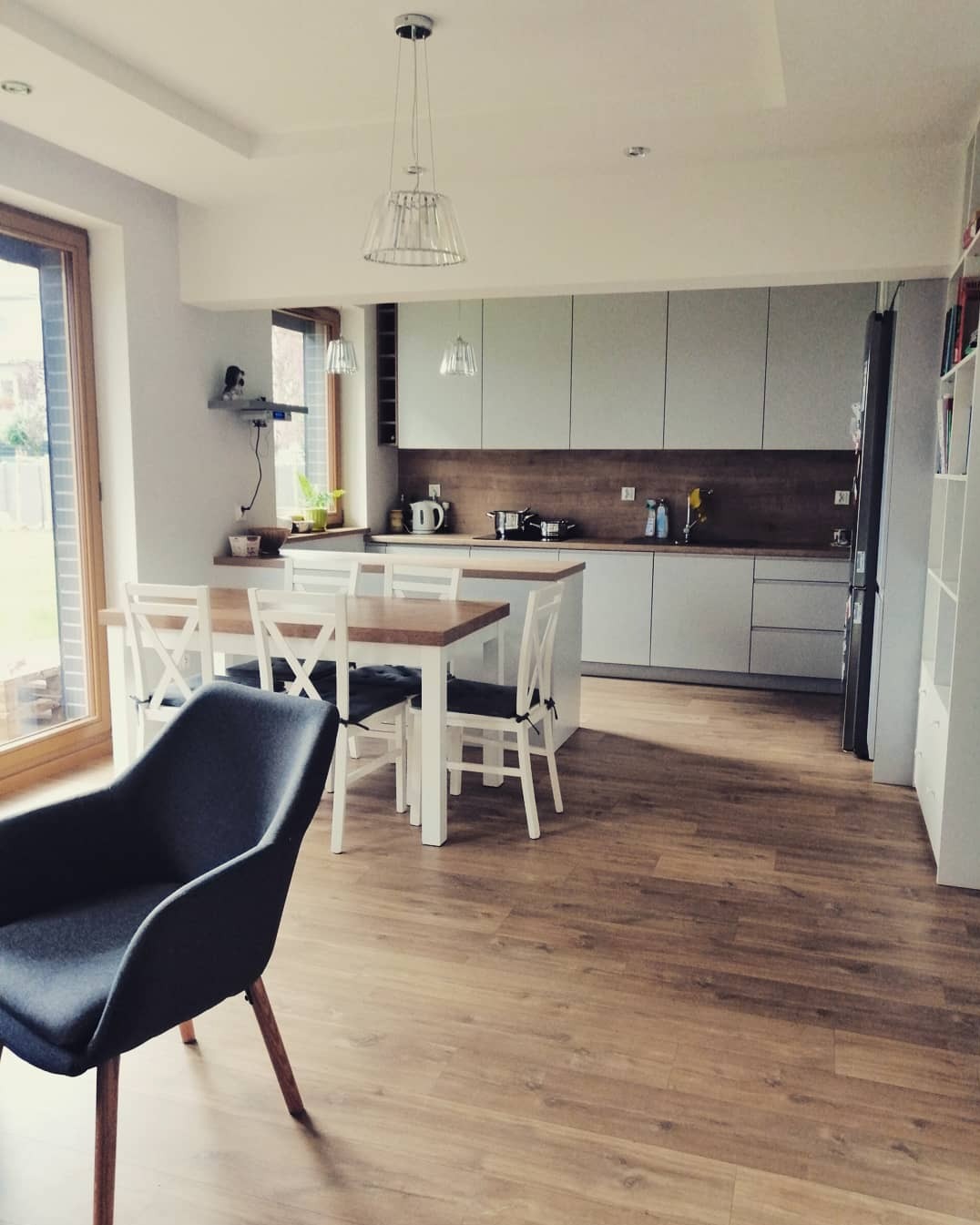 Jasna kuchnia z drewnianym blatem i szarymi szafkami, widoczny stół z białymi krzesłami i ciemnoszary fotel na pierwszym planie, podłoga wyłożona panelami imitującymi drewno.