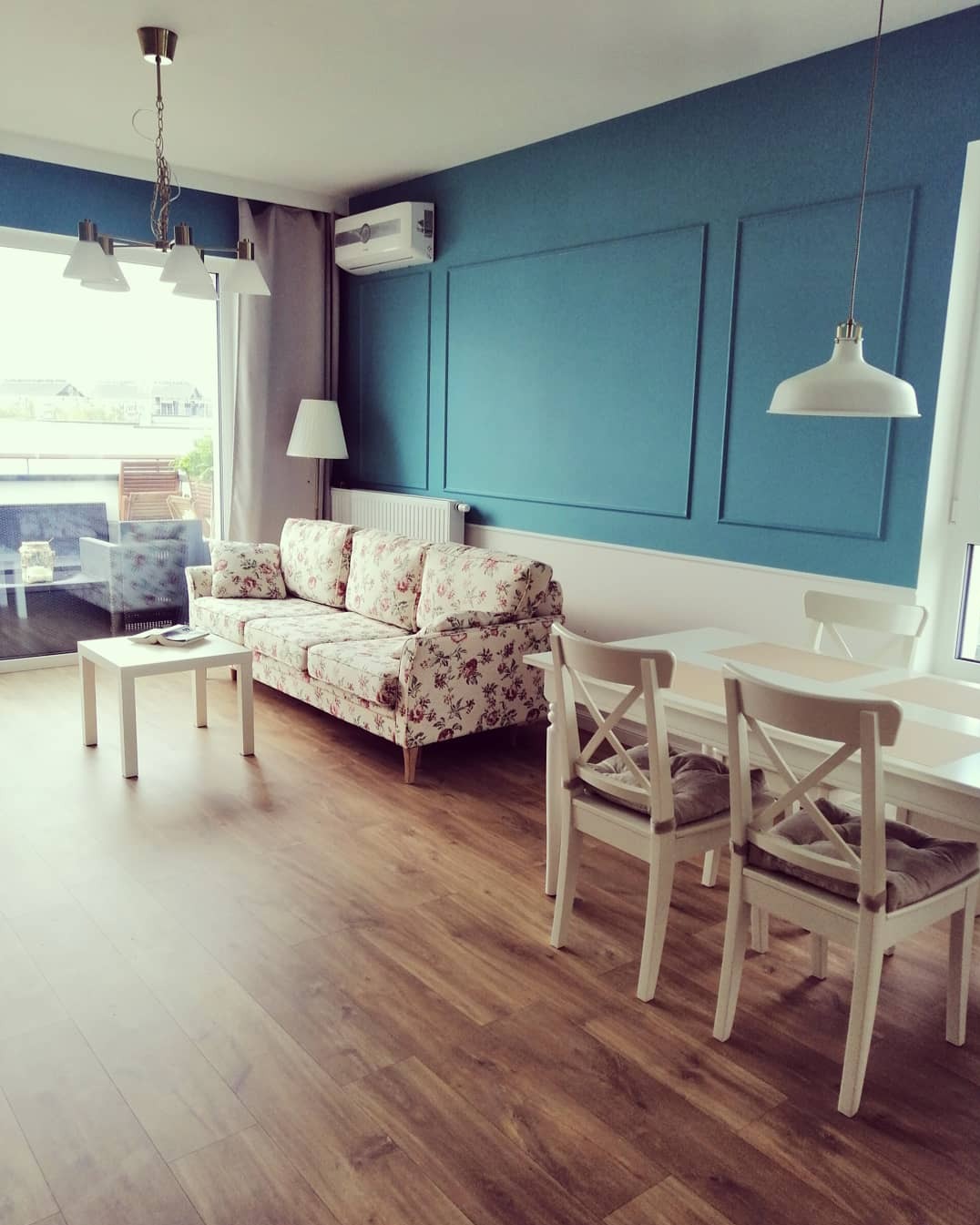 Turkusowa ściana z dekoracyjnymi panelami w salonie, białe meble, wzorzysta sofa i drewnopodobna podłoga.