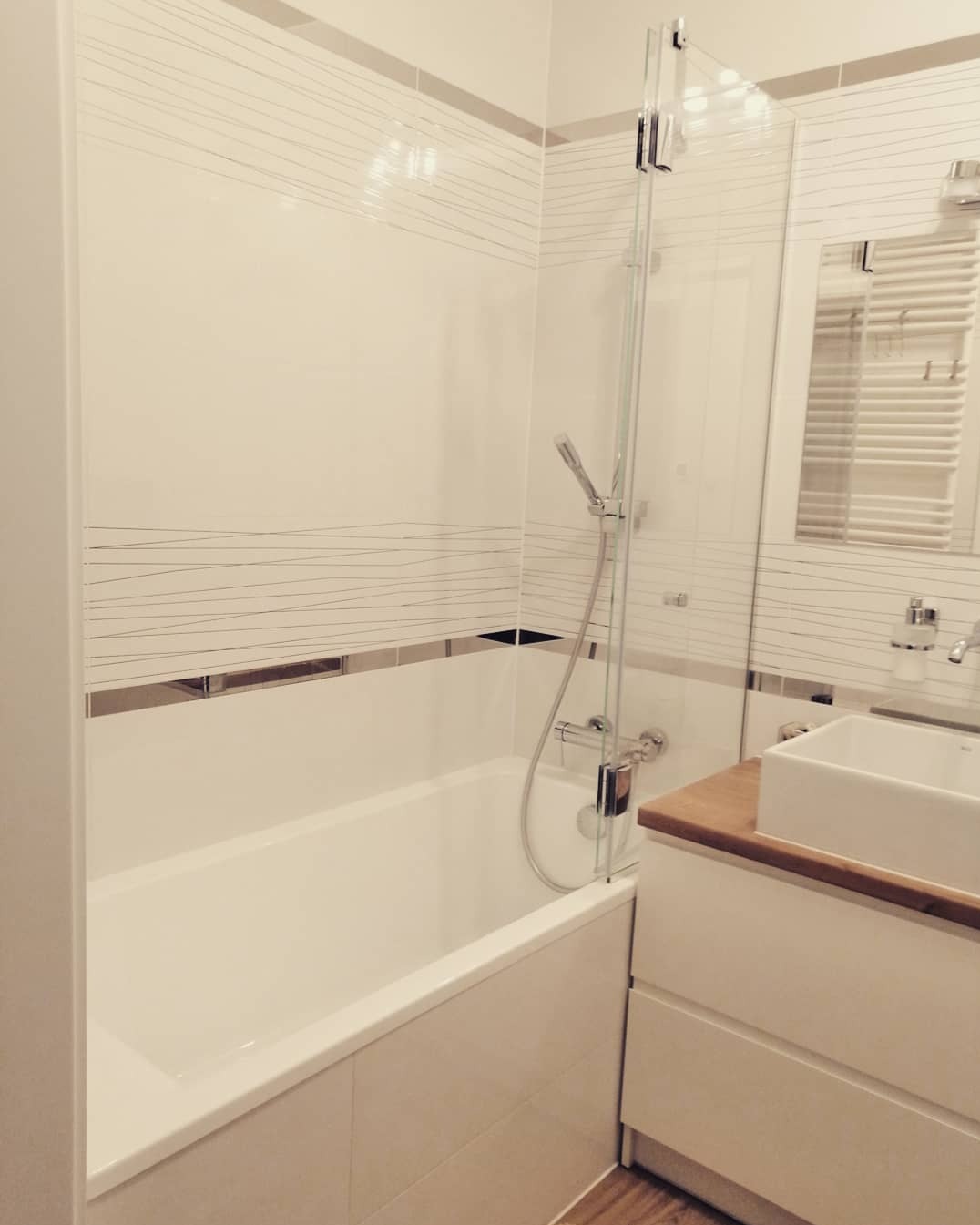 Biała łazienka z wanną i umywalką, wykończona płytkami z delikatnym, poziomym wzorem, kabina prysznicowa z przezroczystego szkła, drewniany blat pod umywalką, minimalistyczny styl.