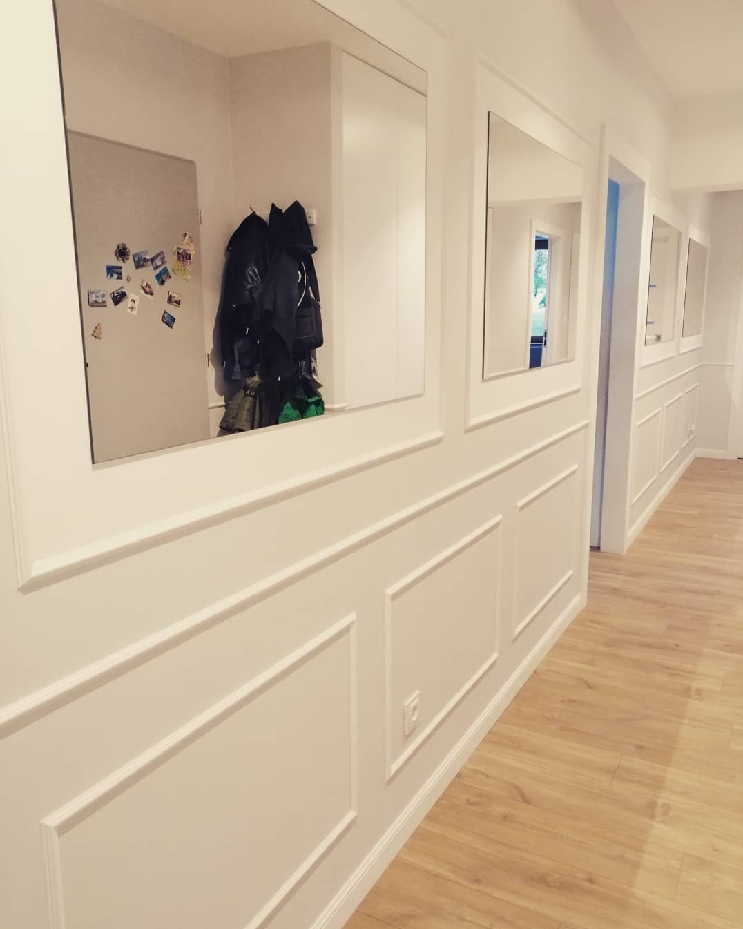 Elegancki korytarz z drewnianą podłogą i białymi ścianami z dekoracyjnymi panelami oraz wbudowanymi lustrami, odbijającymi przedpokój z kurtkami i lodówką z magnesami.